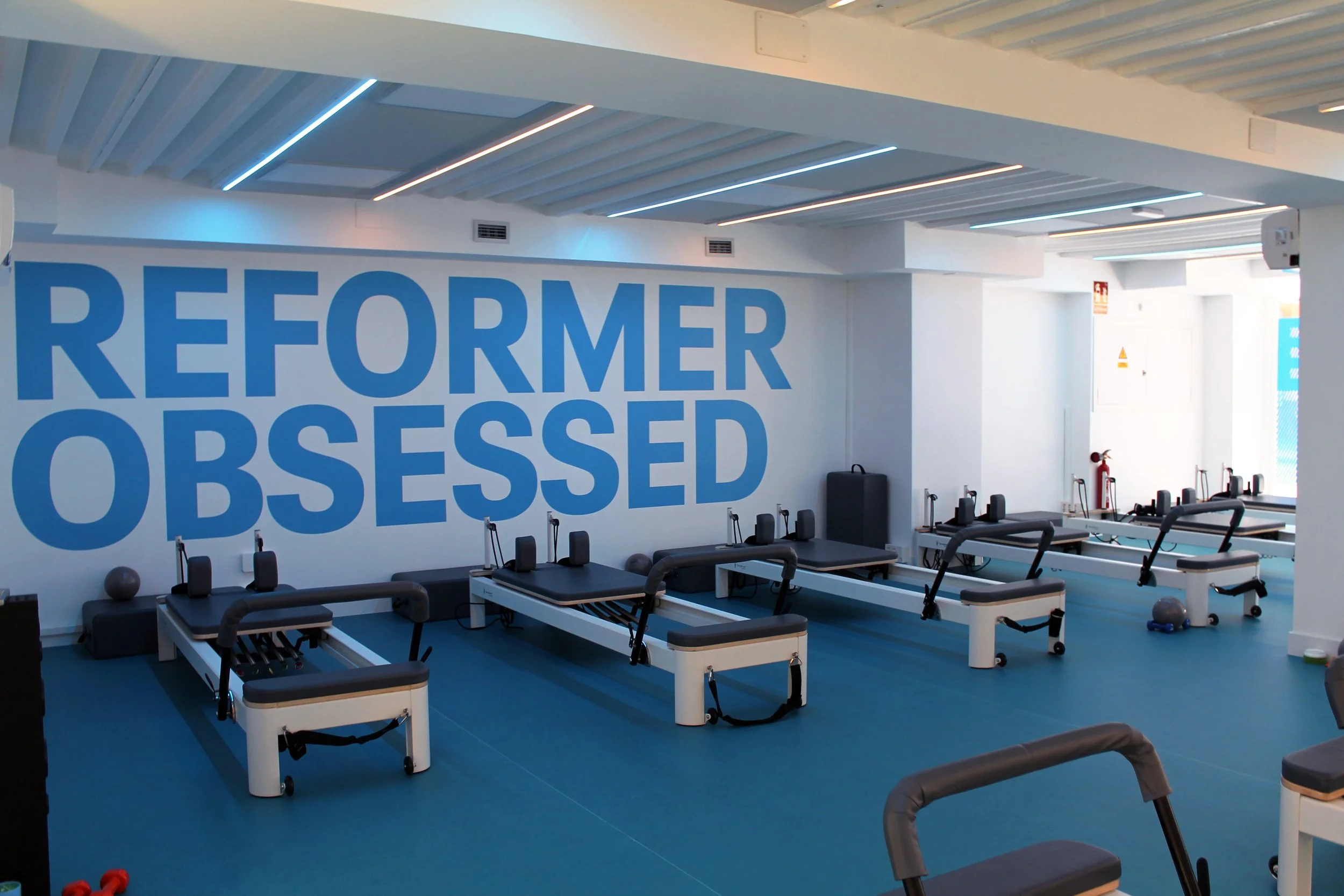 Sala reformer con máquinas de Pilates y etiquetas que dicen 'Reformer Obsessed' en la pared.