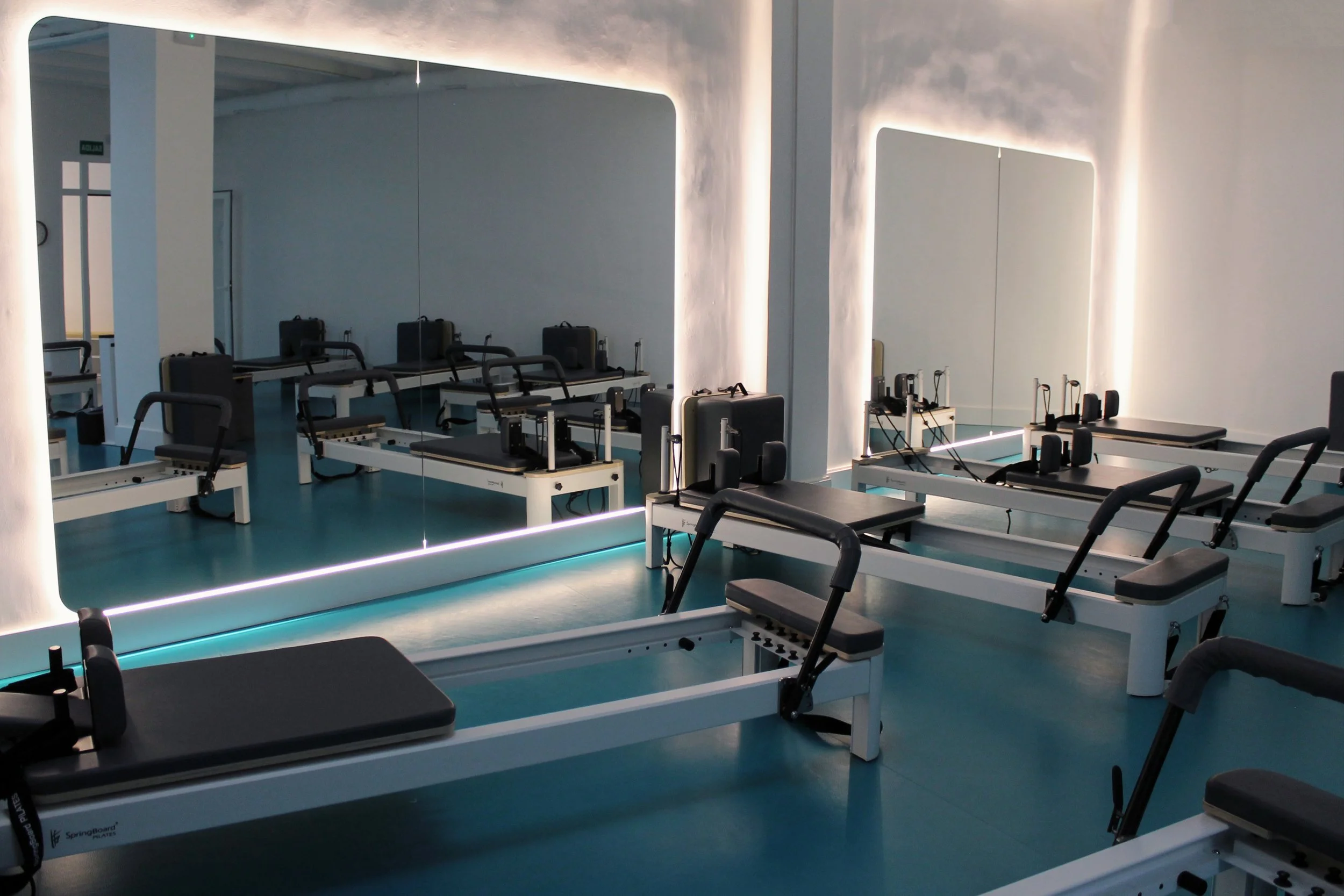 Sala de pilates con varios equipos y espejos que reflejan las máquinas, iluminación moderna y paredes blancas.