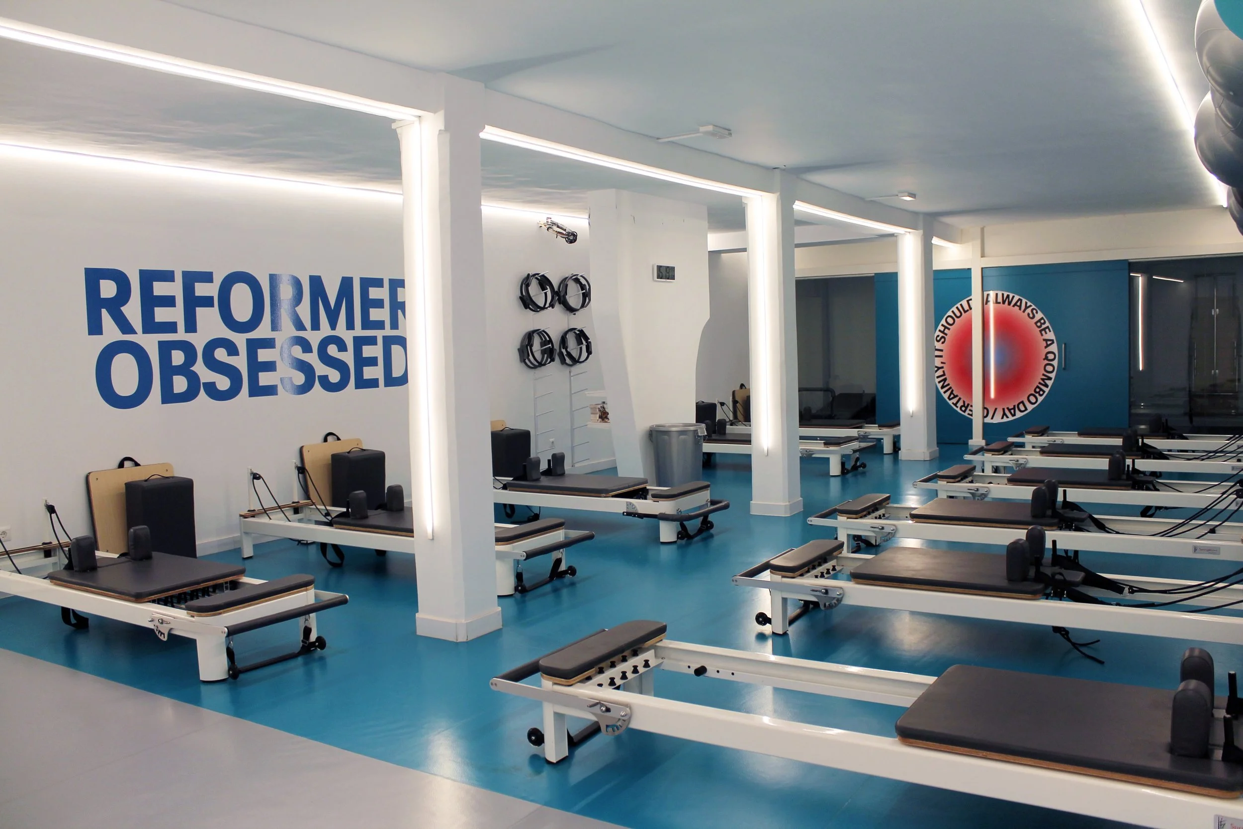 Sala de terapia física con camillas y equipos de ejercicio, pared con cartel que dice 'Reformer Obsession' y decoraciones relacionadas con el ejercicio.