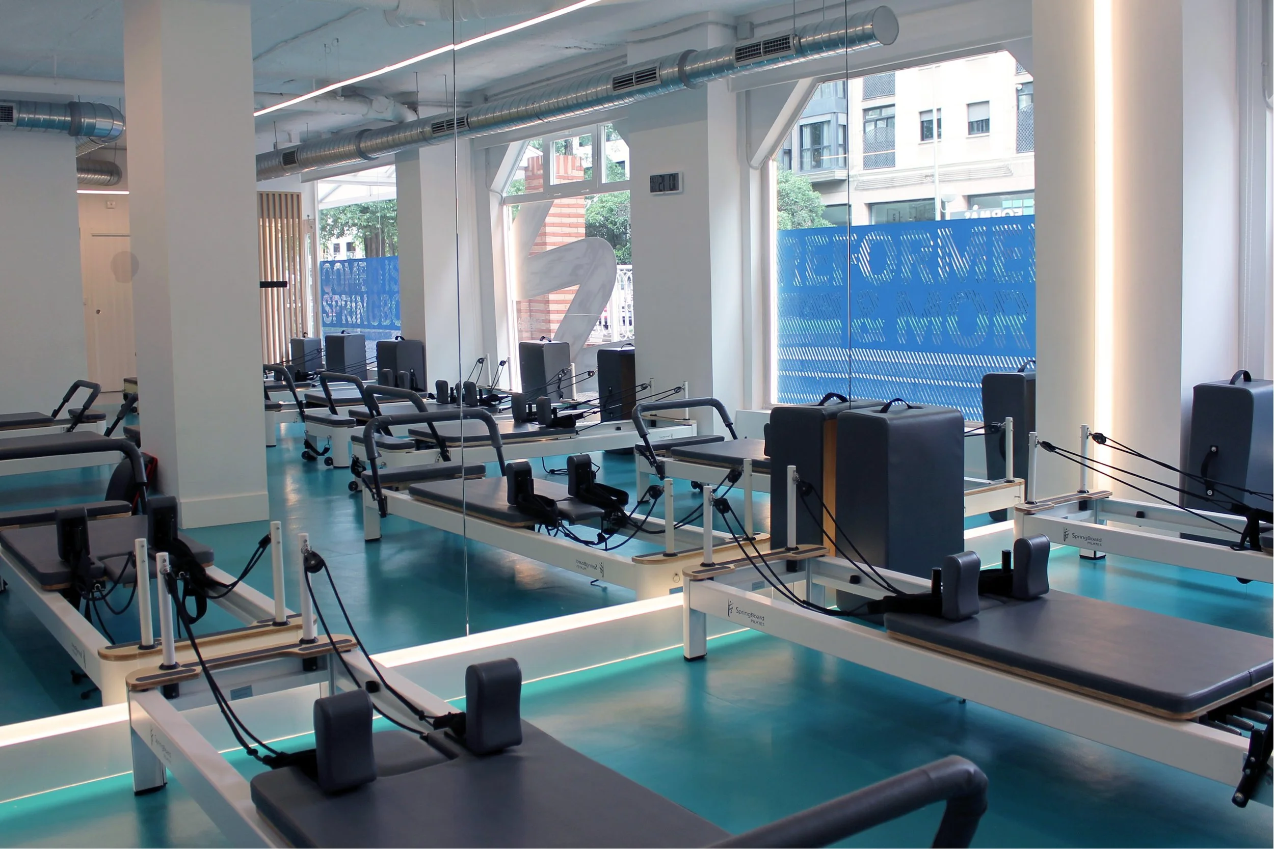 Sala de reformer con varias camas de pilates y máquinas para ejercicios, ventanas grandes y paredes blancas.