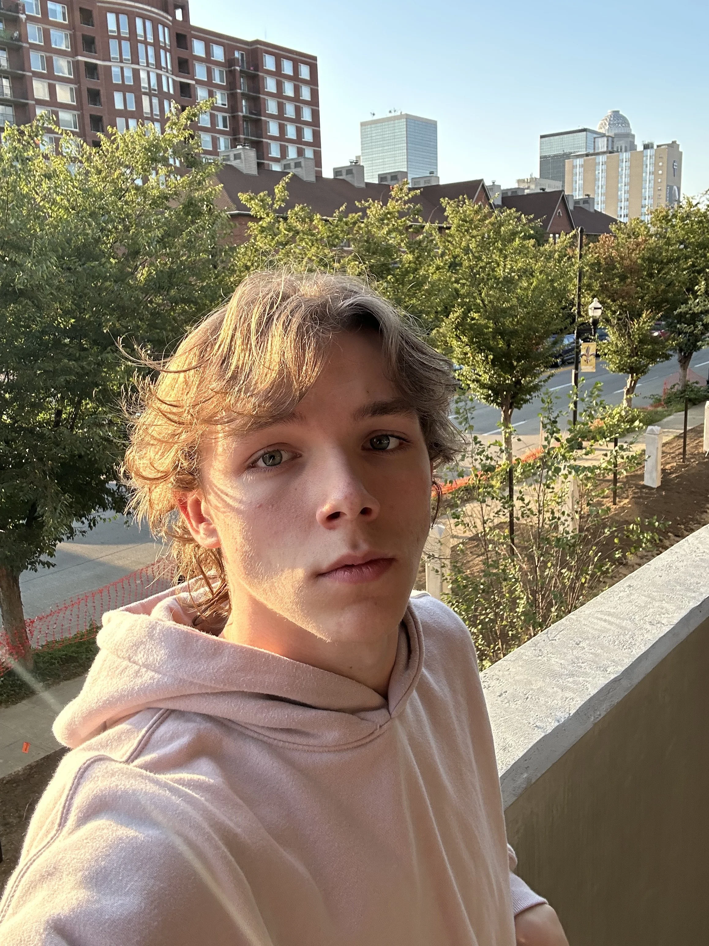 A person with light brown hair and gray eyes taking a selfie on a balcony with a cityscape in the background