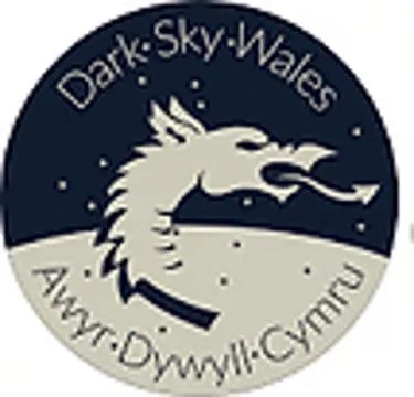 Logo featuring a dragon's head in profile with a background of a moonlit sky and landscape, with text reading "Dark Sky Wales" and "Ayr-Diwyl Cymru".