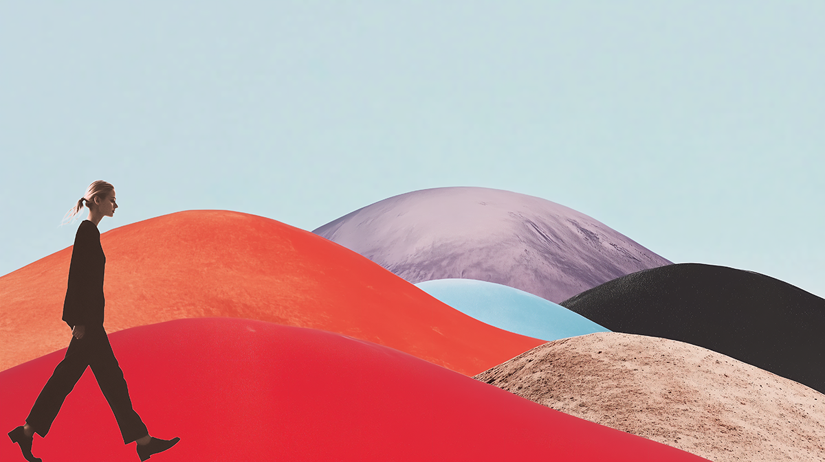A woman walking outdoors with colorful hills or mounds behind her in various shades of red, purple, black, blue, and beige under a clear, light blue sky.