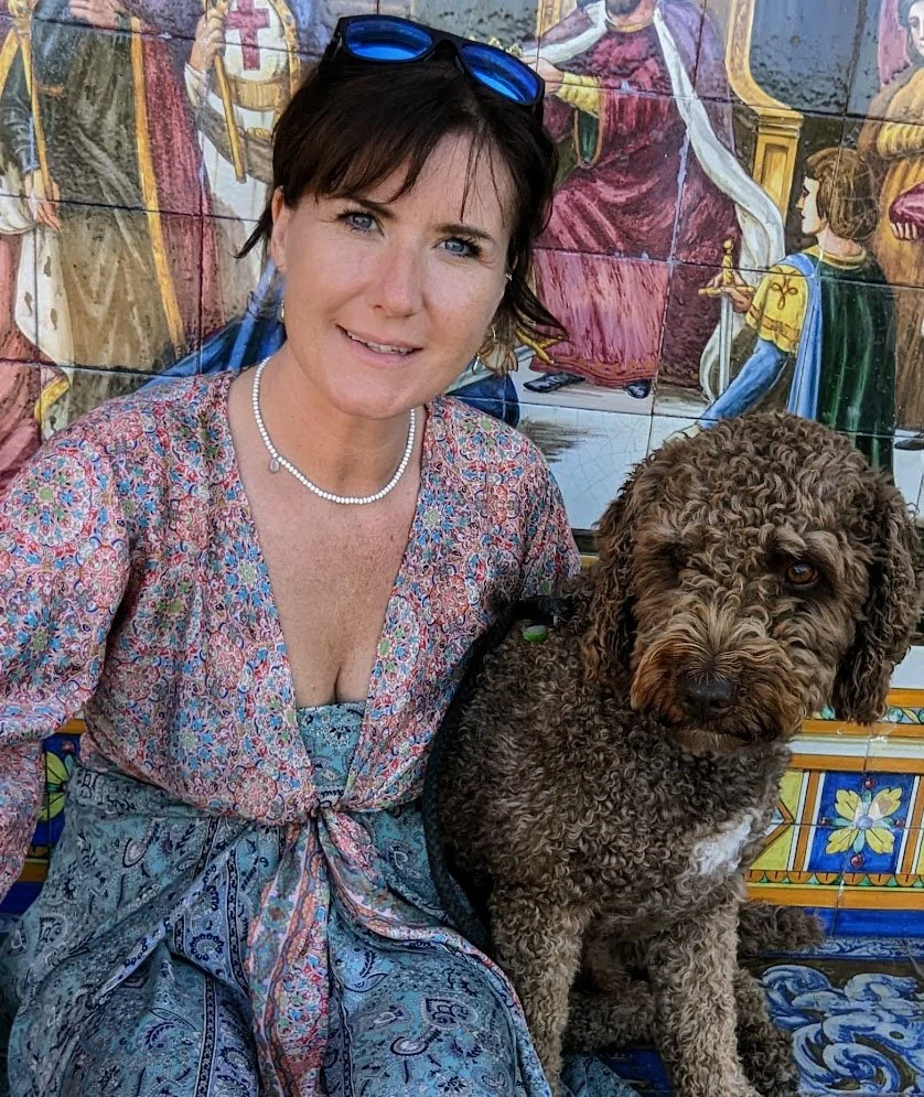 A woman with short dark hair, wearing a floral dress, pearl necklace, and blue sunglasses on her head, sitting next to a curly-haired brown dog with a white patch on its chest against a colorful tiled mural of a religious scene.