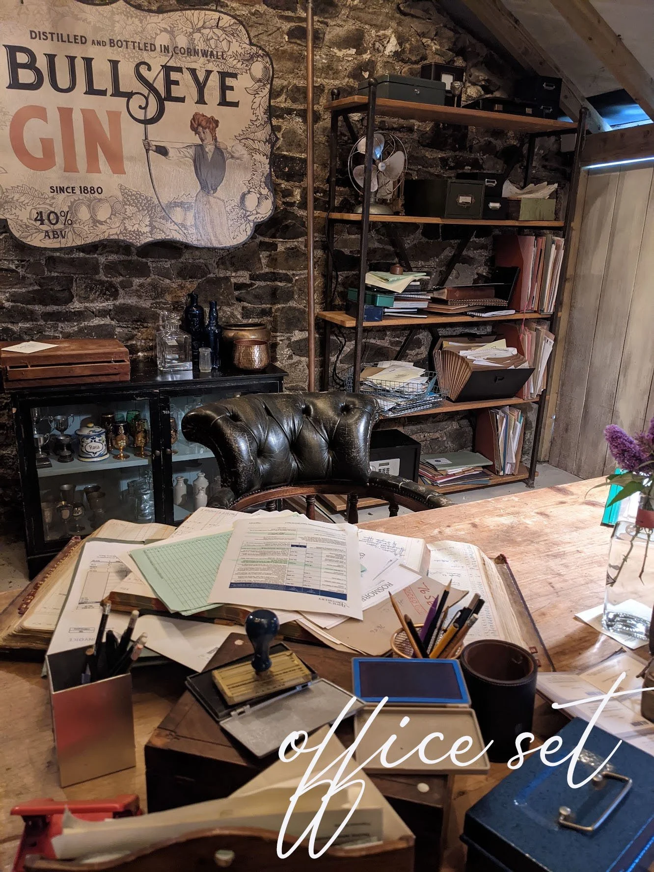 A cluttered office workspace with papers, pens, and office supplies on a wooden desk, a black leather chair, a cabinet with trophies and figurines, a tall bookshelf filled with files and boxes, exposed brick wall, and a vintage-style sign for Bullseye Gin hanging on the wall.