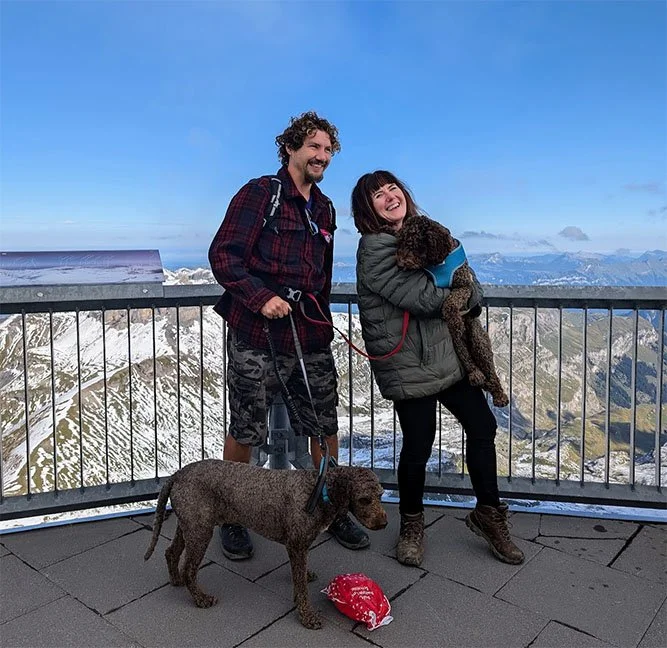 Two people smiling on an outdoor observation deck with snowy mountains in the background. One person is holding a dog, and a second dog is on the ground with a red bandana nearby.