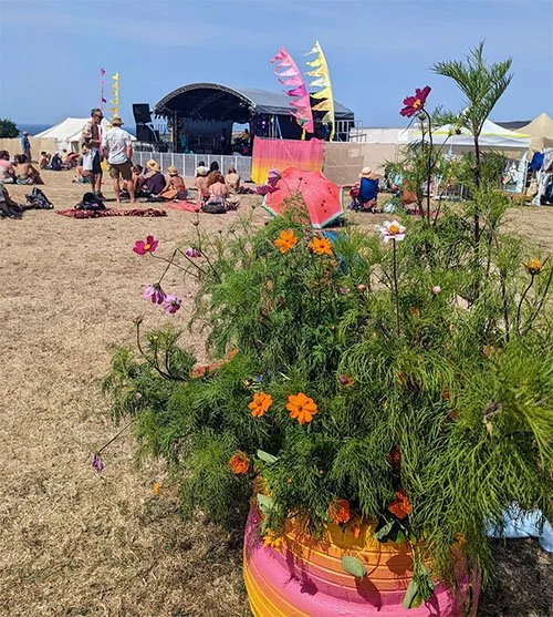 Flower arrangement in a colorful pot with a beach scene, stage, and people sitting on the sand in the background
