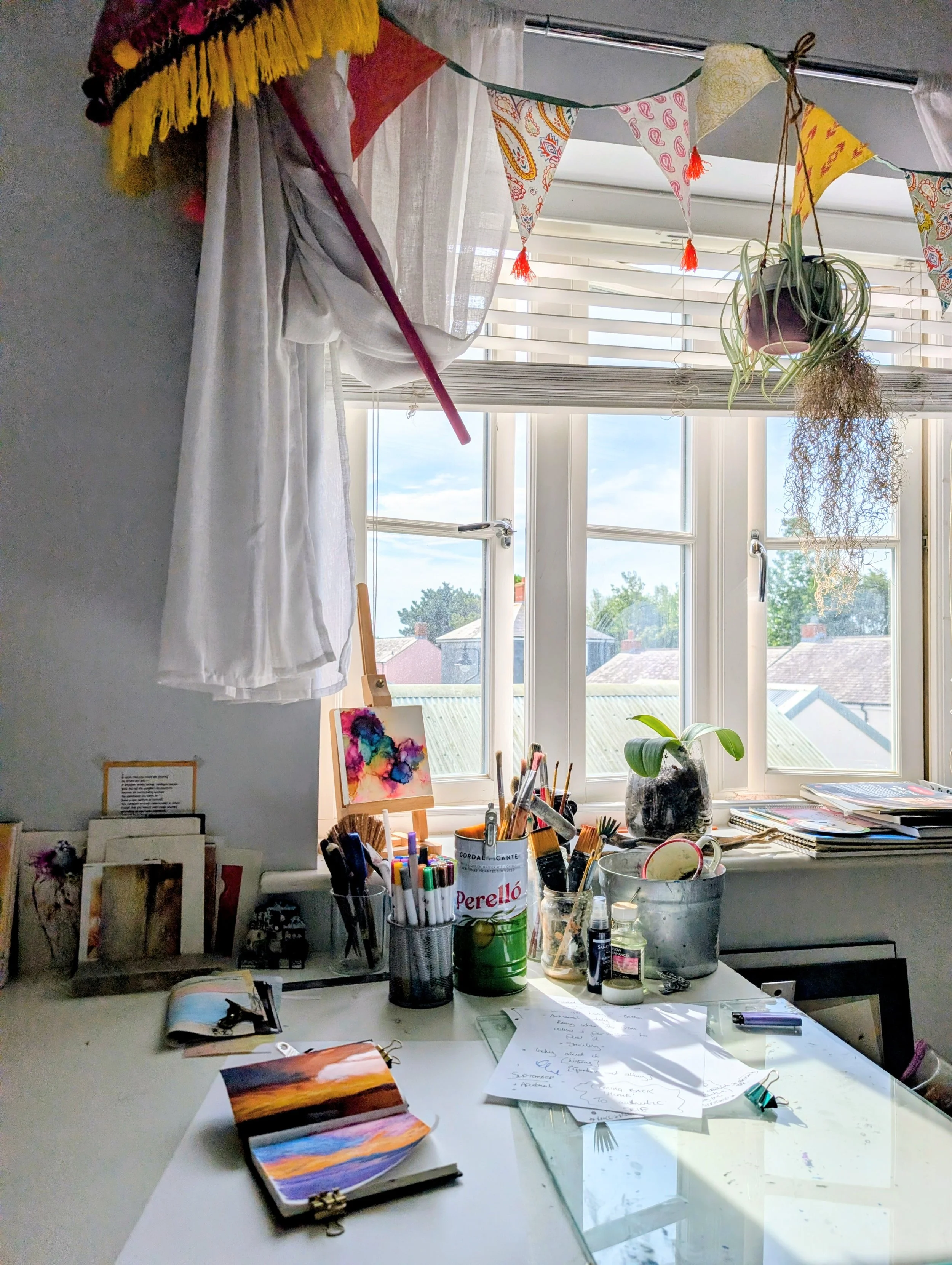 A cluttered art workspace with a desk and window. The desk has painted notebooks, art supplies including pens, brushes, and jars. Hanging on the window are colorful decorative flags. The window shows a view of rooftops and trees under a sky.