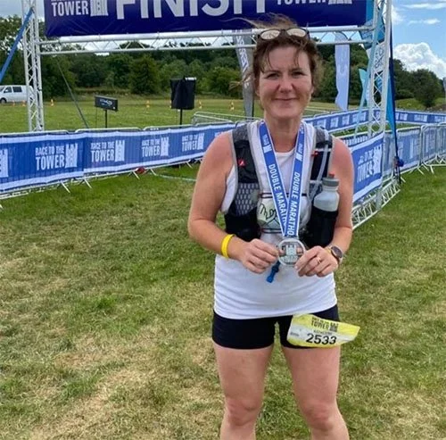 A woman standing at a race finish line holding a medal, wearing a race bib, wristband, and hydration pack, in an outdoor grassy area with trees and a clear sky in the background.