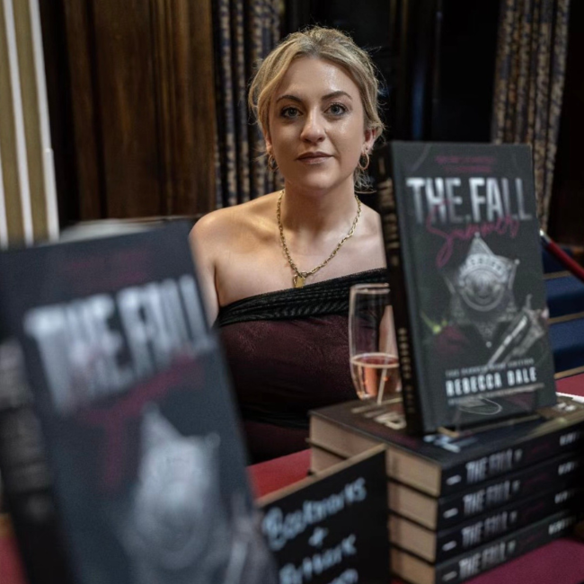A woman with blonde hair, wearing a black strapless top and gold necklace, sitting at a table with multiple copies of a book titled 'The Fall' by Roderic Eale, and a glass of pink wine.