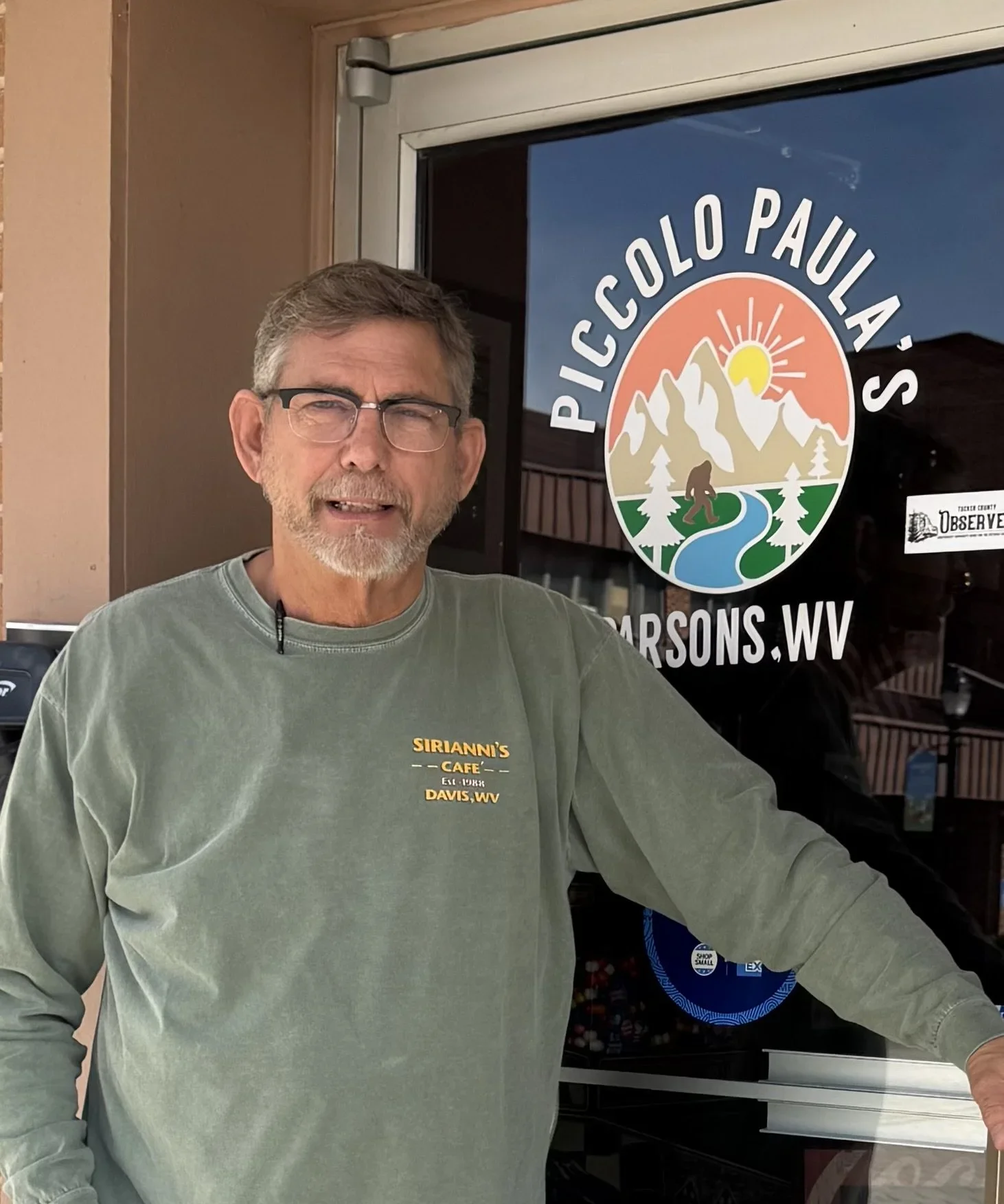 A man with glasses and a green long-sleeve shirt standing in front of a door with a sign that reads 'Piccolo Paula's' in Marsons, West Virginia, with mountains, pine trees, and a setting sun depicted on the sign.