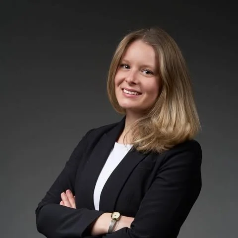 A woman with shoulder-length blonde hair, wearing a black blazer and white top, smiling with arms crossed against a dark gradient background.