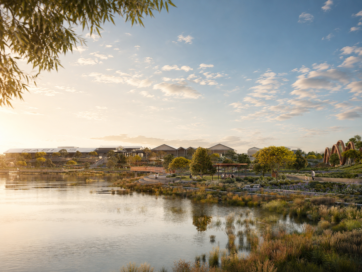 A scenic view of a park by a lake during sunset with trees, a walking path, and modern buildings in the background.