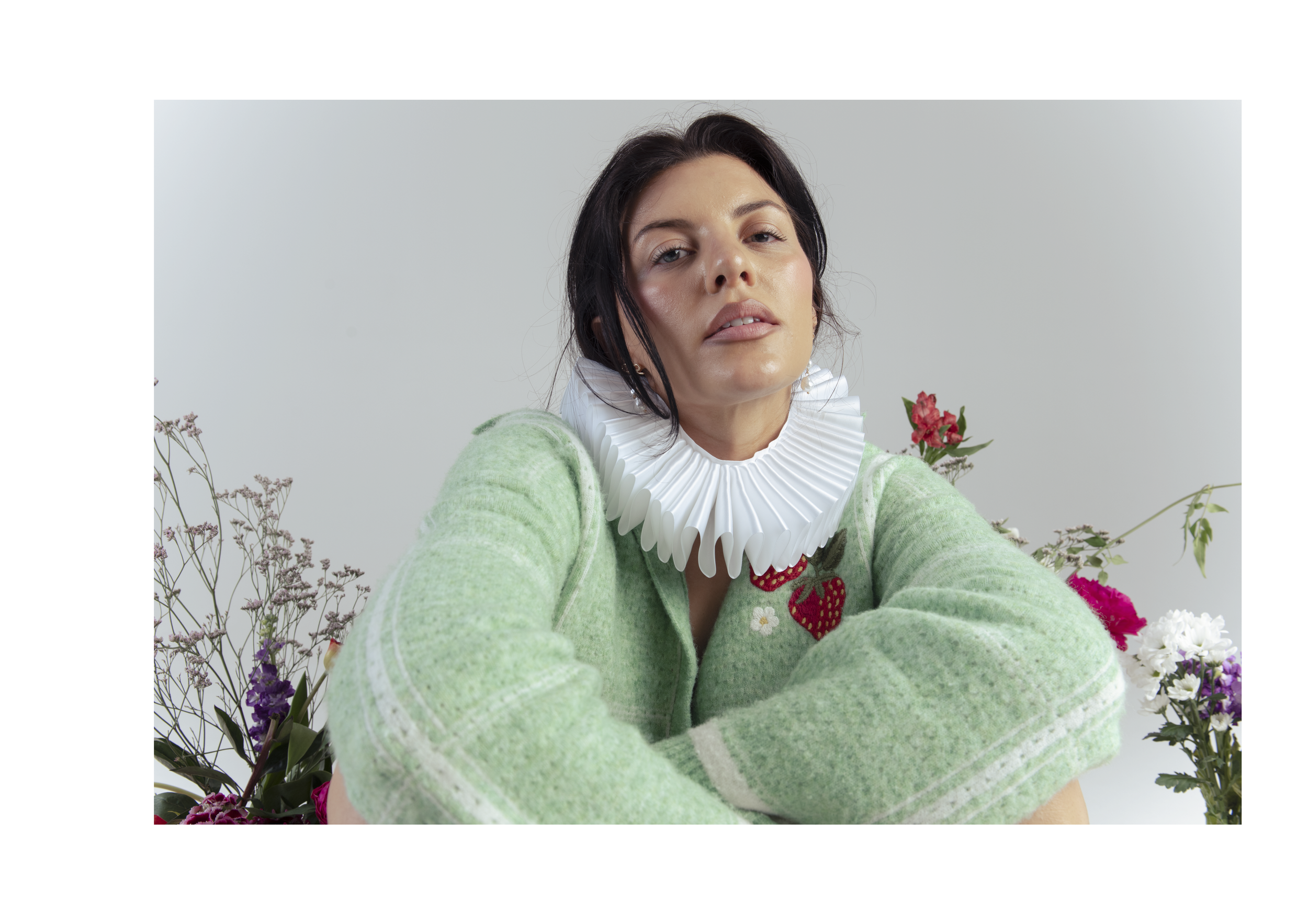 A woman with dark hair and fair skin wearing a green sweater with strawberry and flower designs, and a white ruffled collar, is surrounded by various colorful flowers against a plain light gray background.