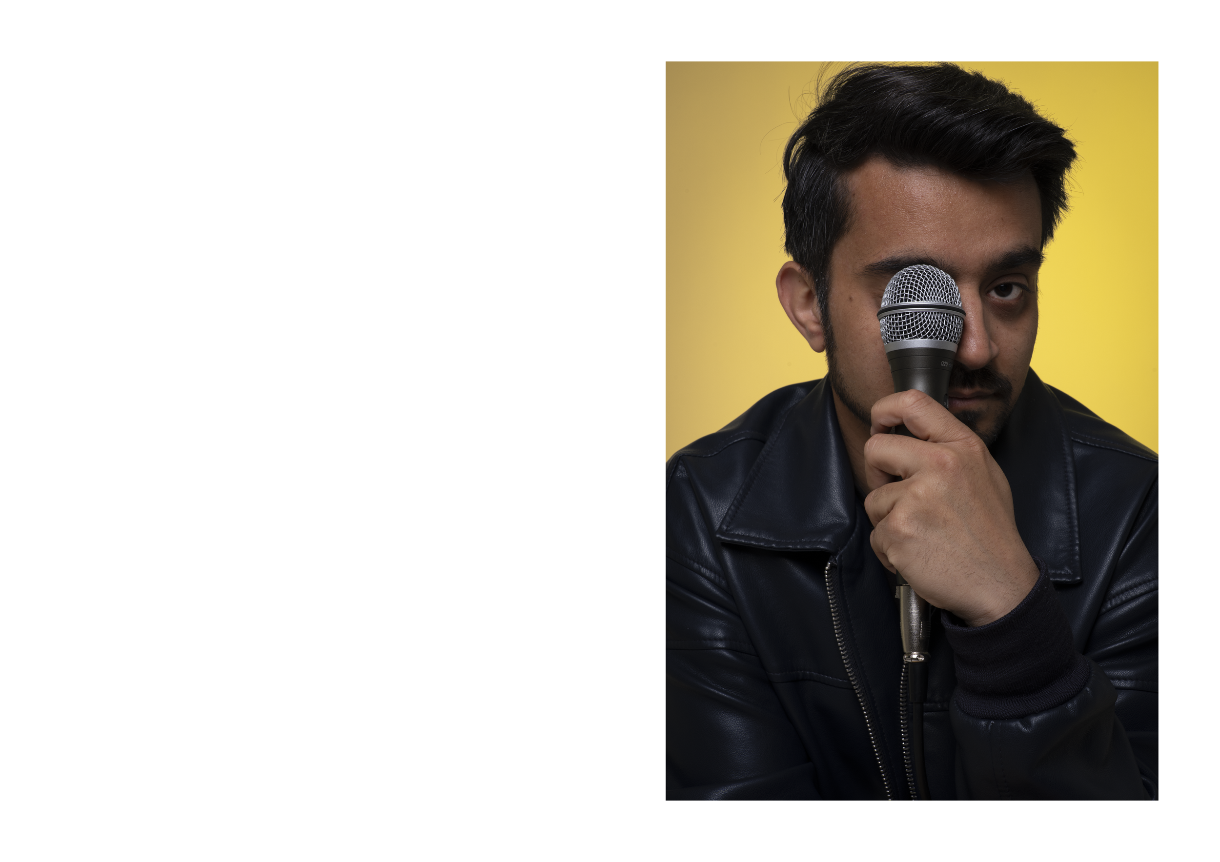 A young man with dark hair and a beard holding a microphone close to his face, wearing a black leather jacket, with a yellow background.