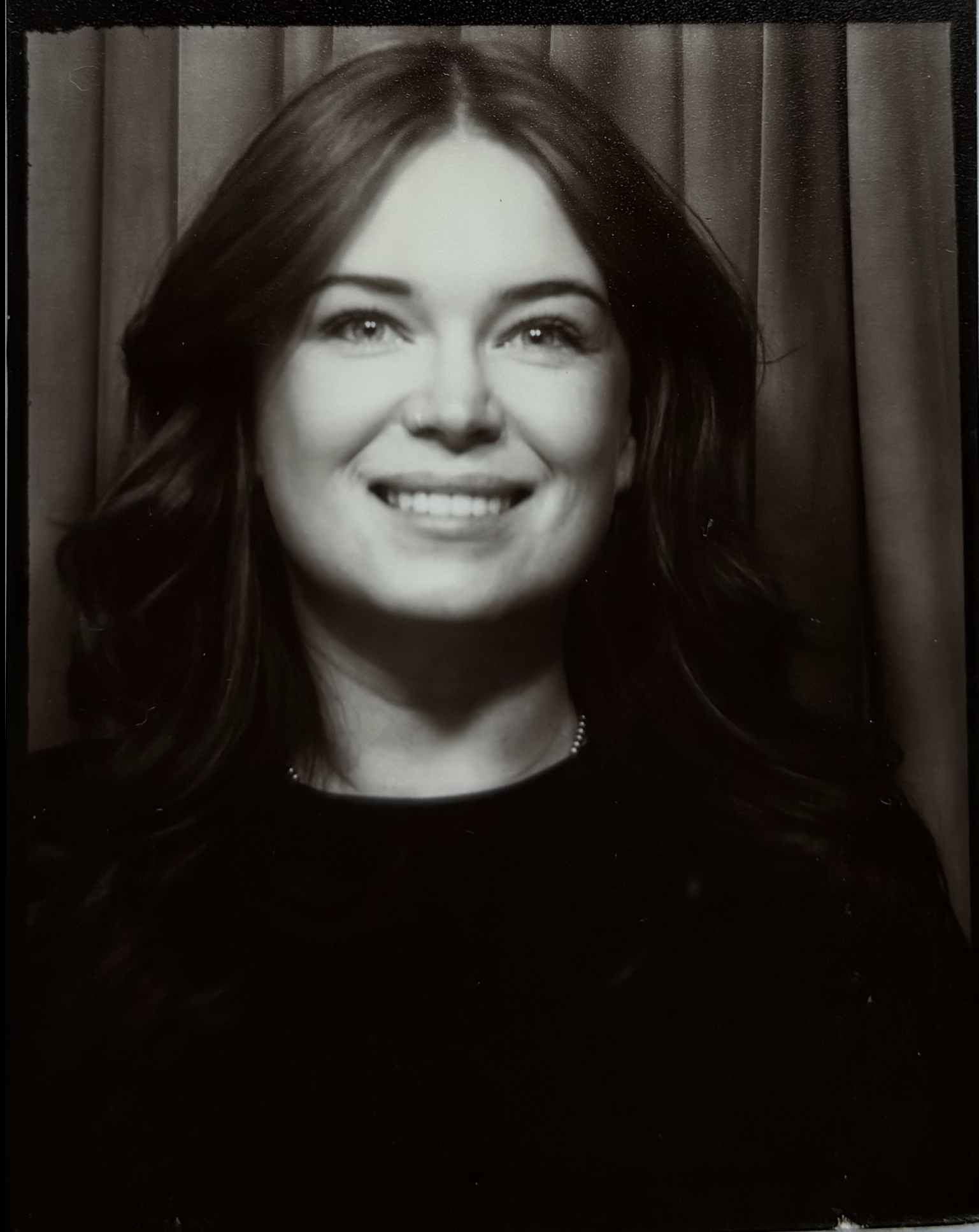 Black and white portrait of a woman with long dark hair, smiling, with curtains in the background.