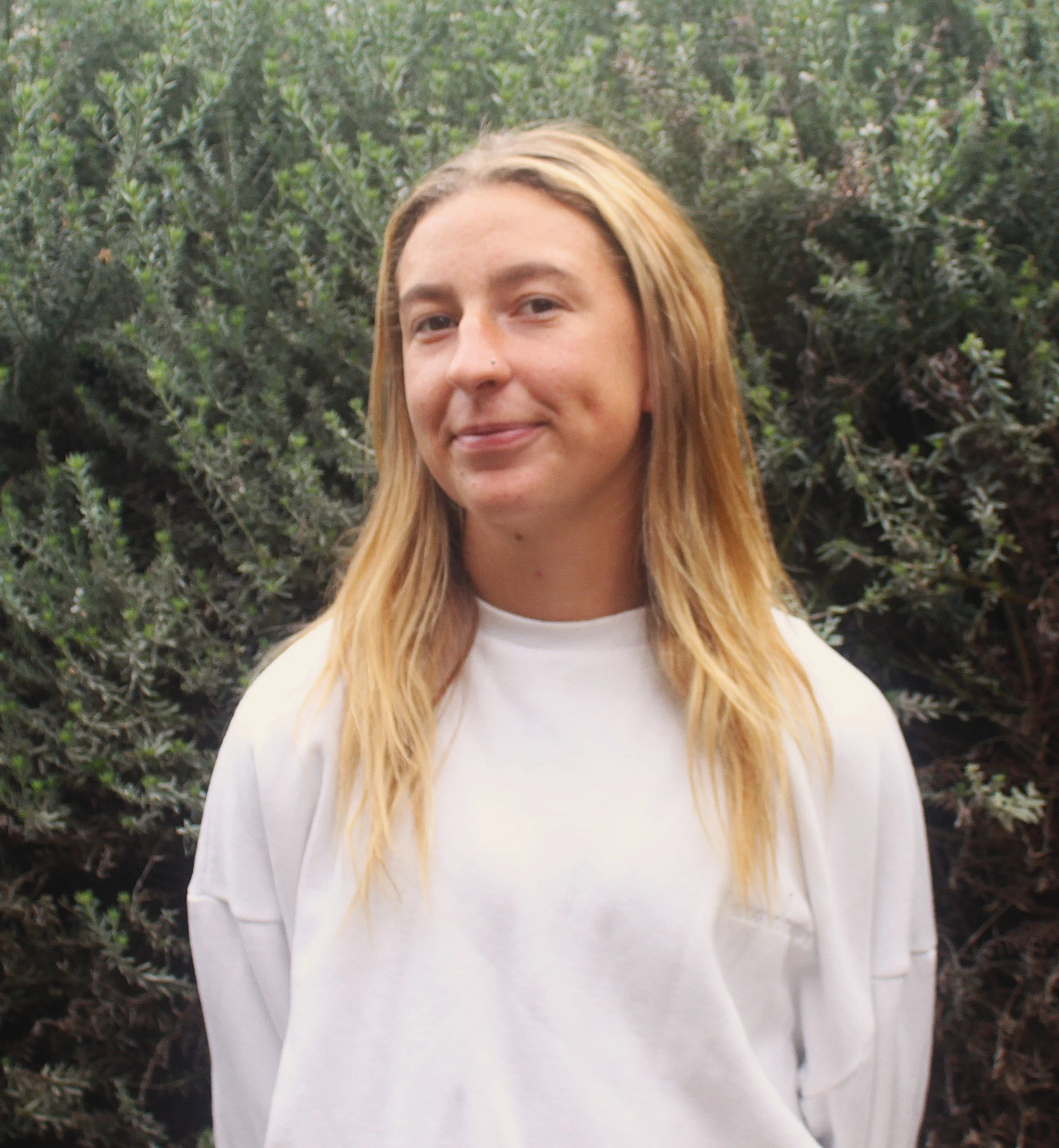 A woman with long blonde hair, wearing a white shirt, stands outdoors with greenery in the background.