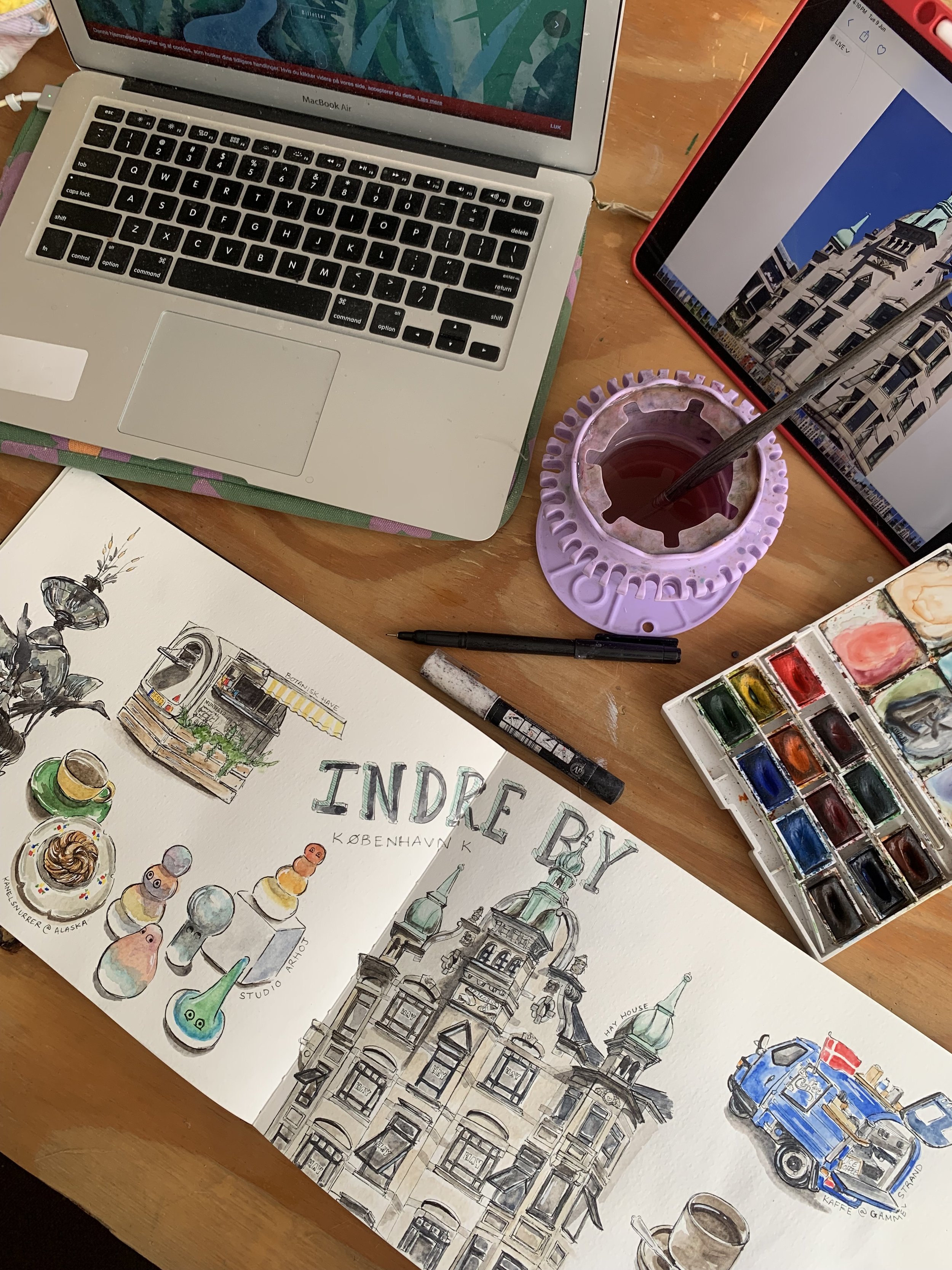 A workspace with a MacBook Air laptop, an iPad or tablet, a purple cup with tea and a spoon, a watercolor paint set, and a sketchbook featuring watercolor illustrations of buildings, coffee cups, and sketches of European architecture.