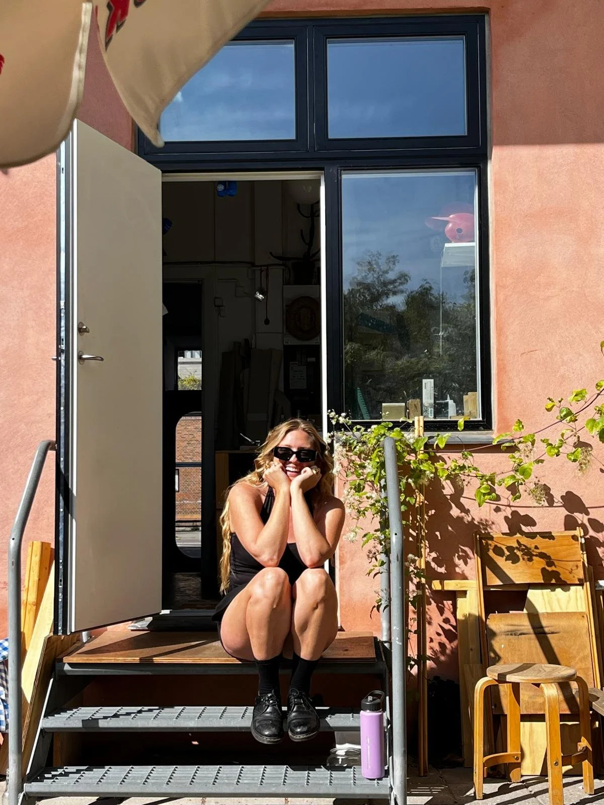 A woman with long blonde hair, wearing black sunglasses, a black tank top, black shorts, and black boots, sits on the steps of an open door of a pink building. She is smiling and resting her chin on her hands. There is a wooden chair and a pink water bottle beside her. Sunlight casts shadows of the plant on the wall.