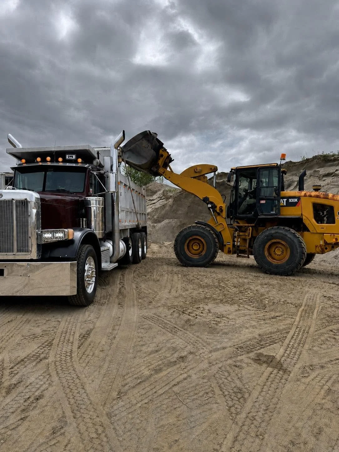 Construction site with a large yellow front loader transferring dirt or sand into a black and maroon dump truck under a cloudy sky.