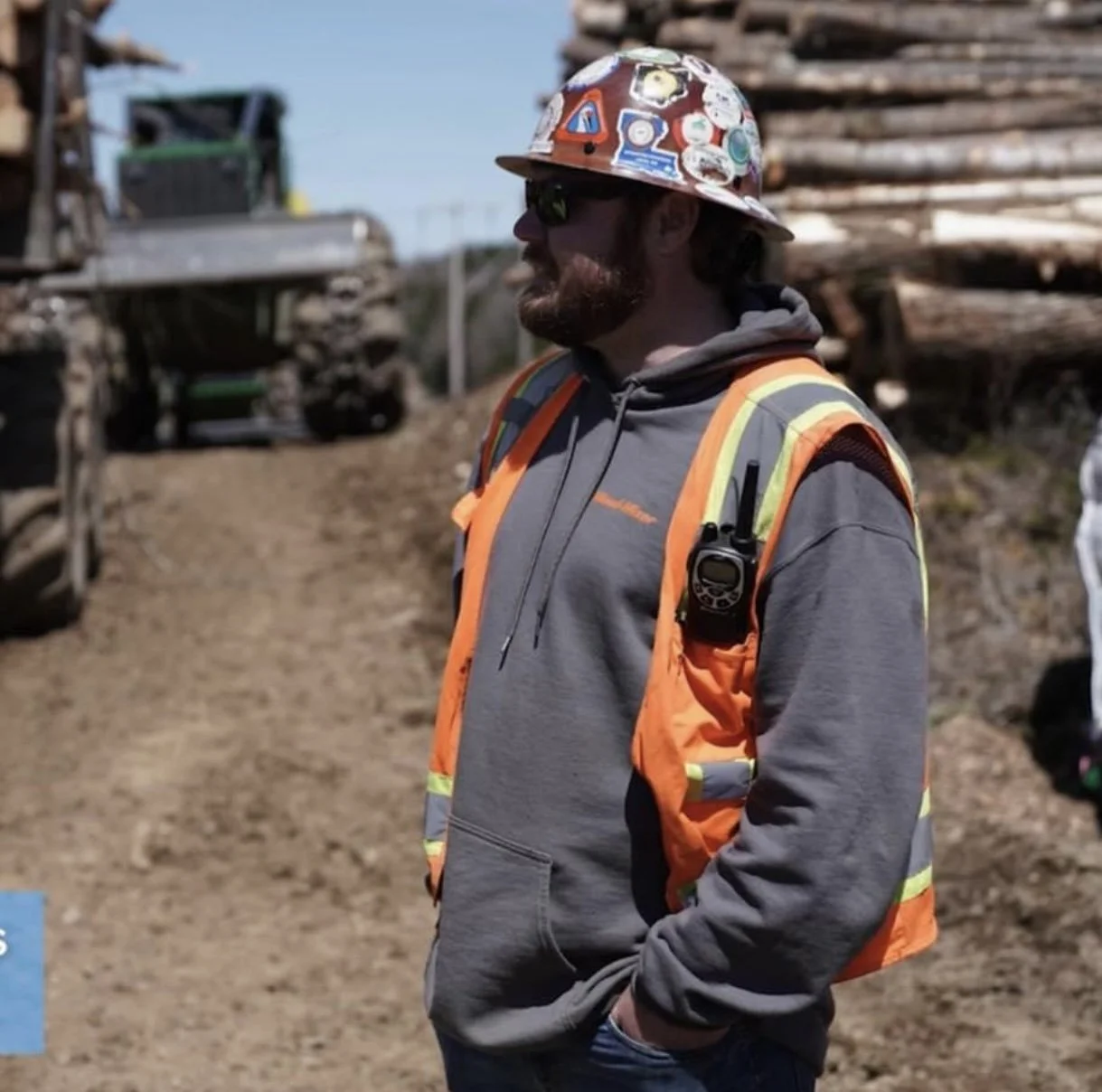 A construction worker wearing a gray hoodie, orange safety vest, and a colorful safety helmet with stickers, standing outdoors at a construction site with logs and machinery in the background.