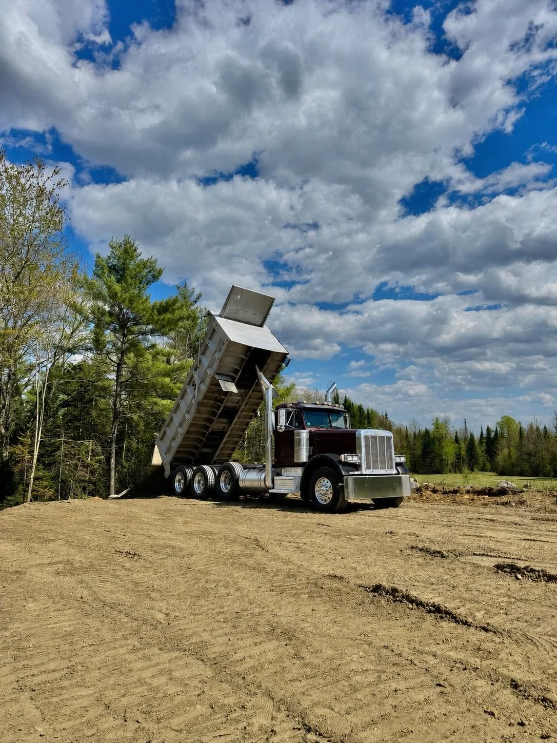 A dump truck with a black cab and silver chassis on a dirt field, with its bed raised spilling dirt, under a partly cloudy sky, with green trees in the background.