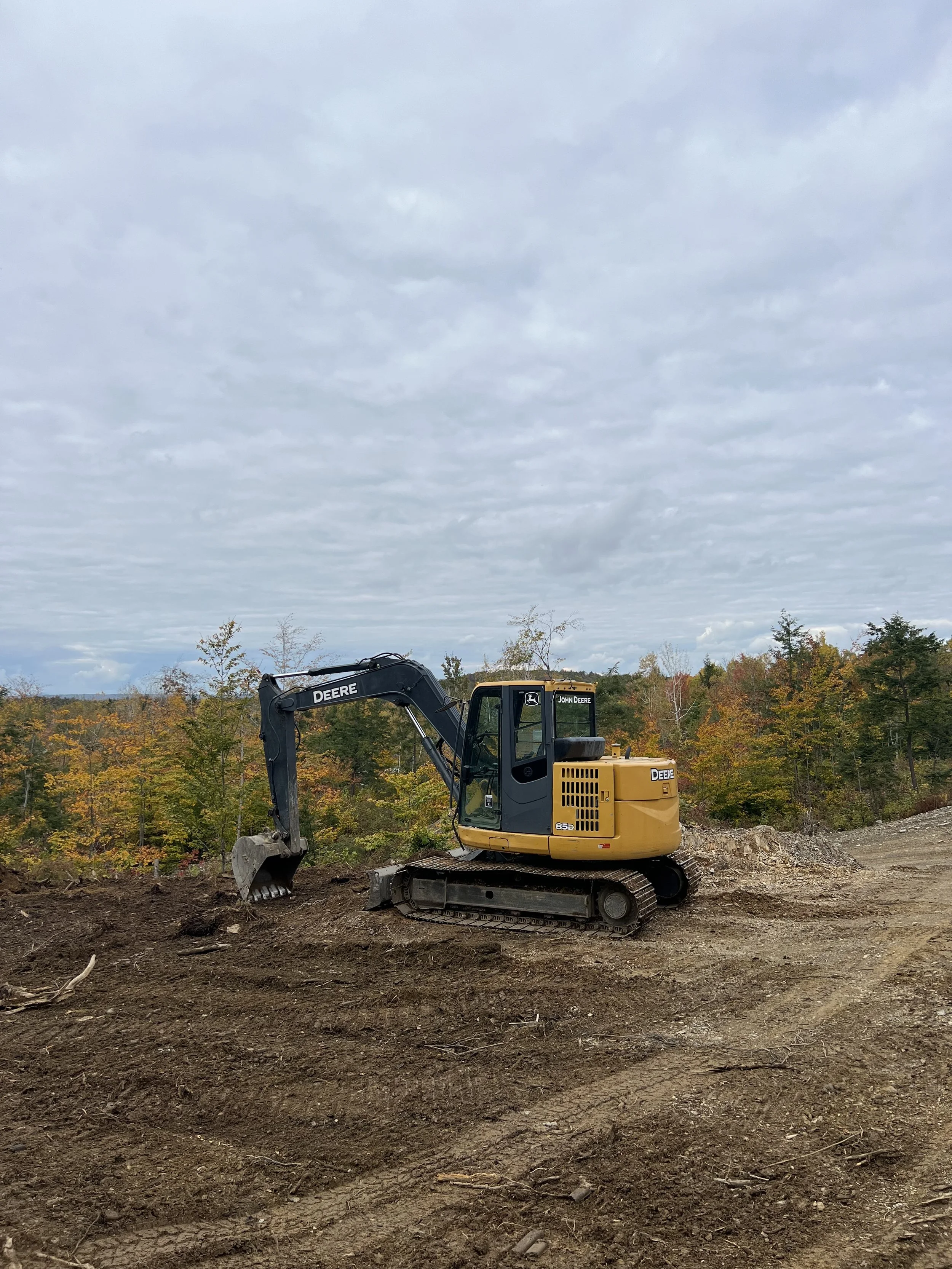 A yellow John Deere excavator working on a dirt construction site, with trees in autumn colors and a cloudy sky in the background.