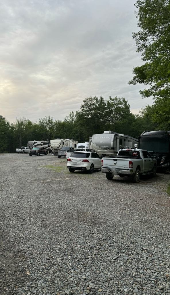 A gravel parking lot with several parked SUVs and large RVs, set against a backdrop of trees and a cloudy sky.