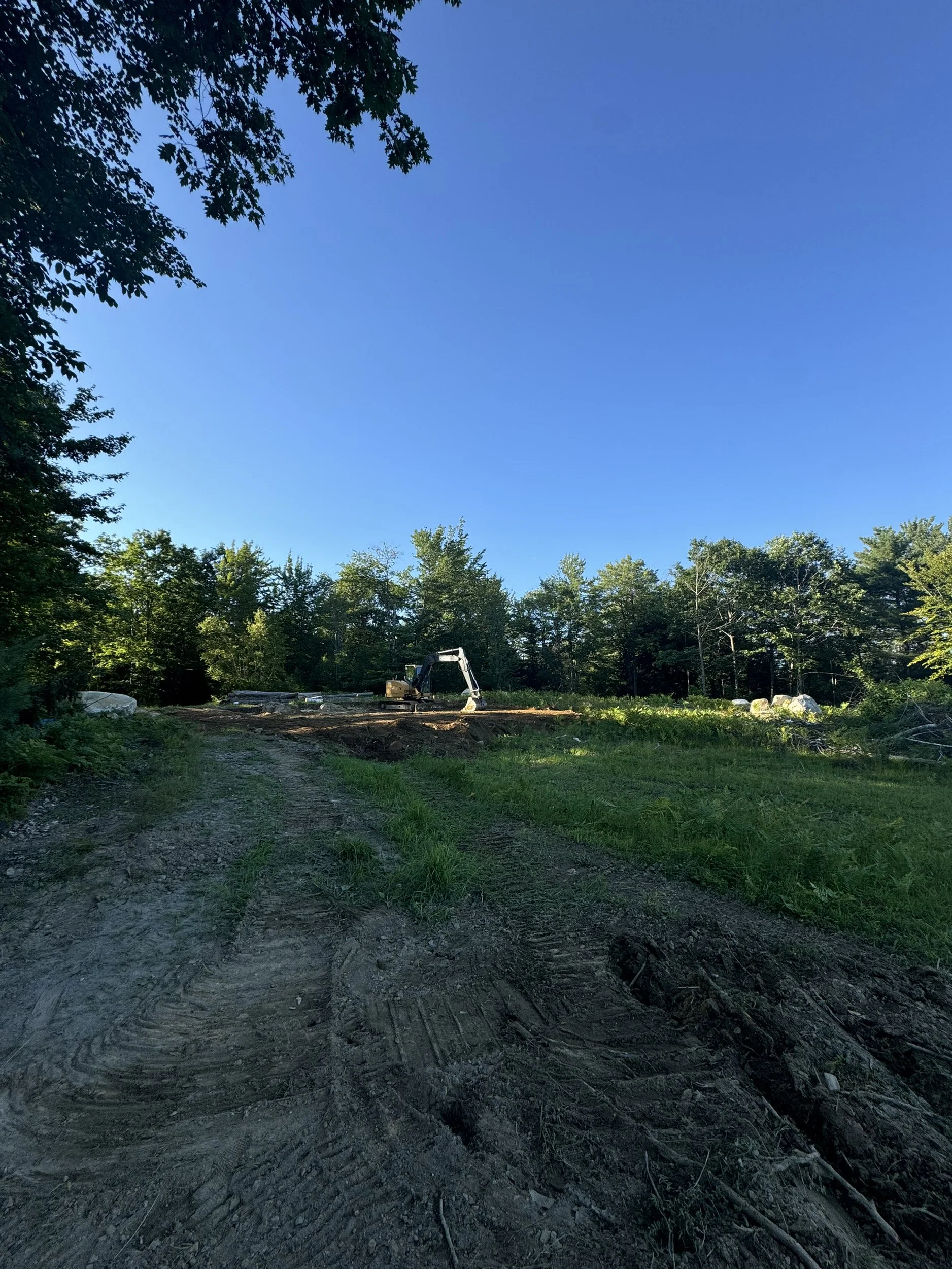 A dirt clearing in a wooded area with a small excavator and tire tracks, surrounded by trees under a clear blue sky.