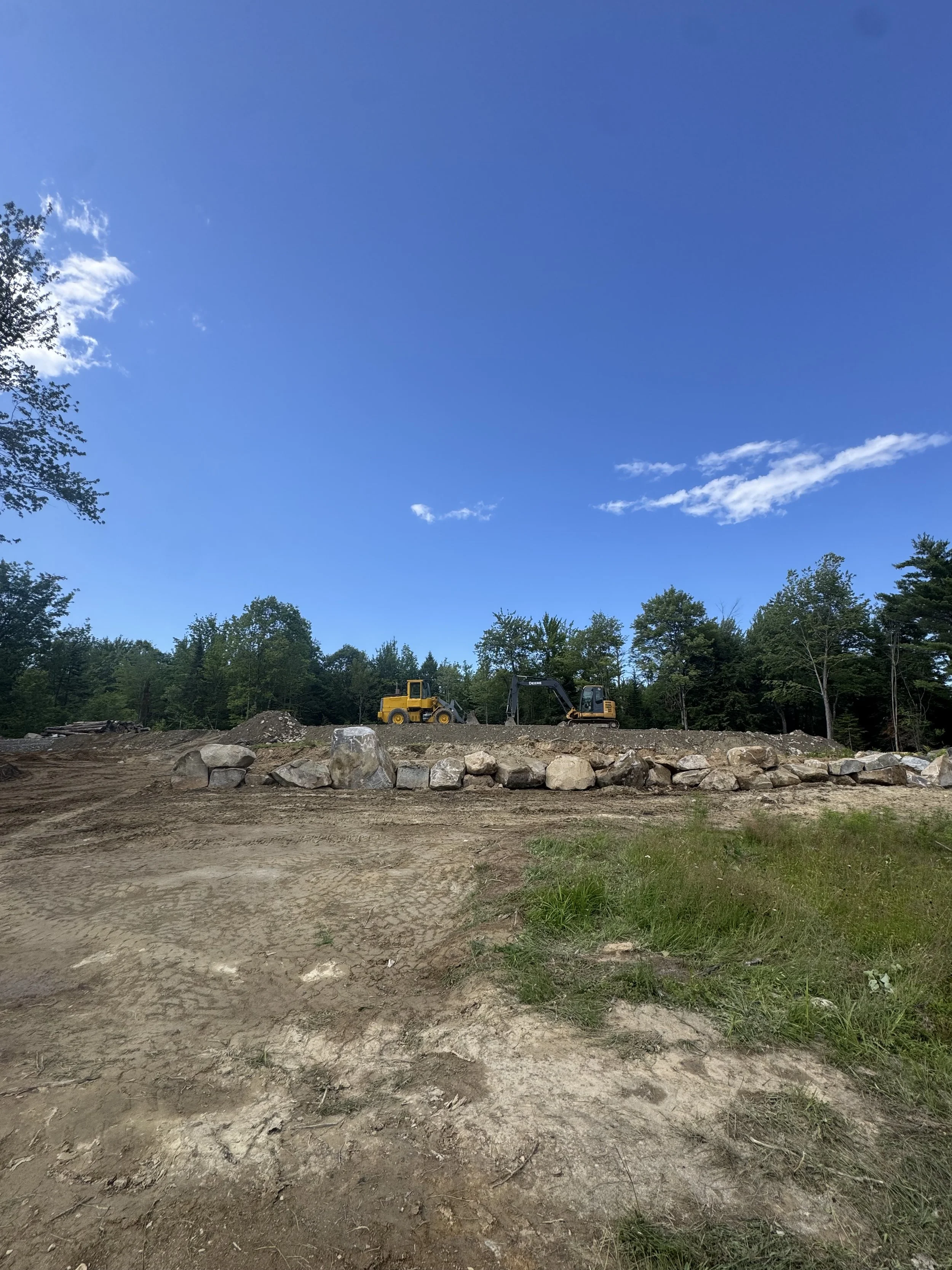 Construction site with two small yellow excavators and piles of rocks, surrounded by trees under a clear blue sky with a few clouds.