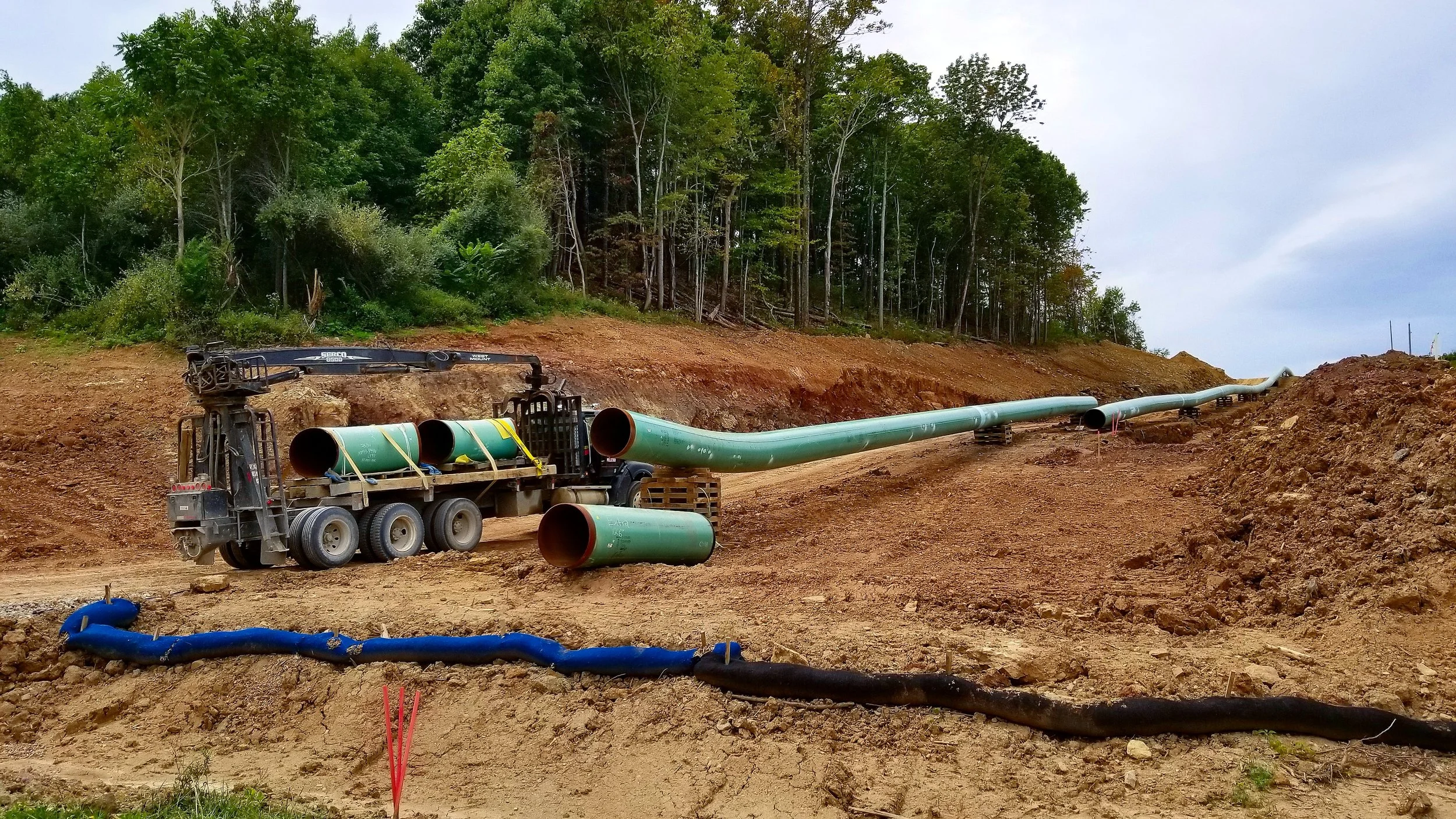 Construction site with green pipelines being laid in a dug area surrounded by dirt and trees.