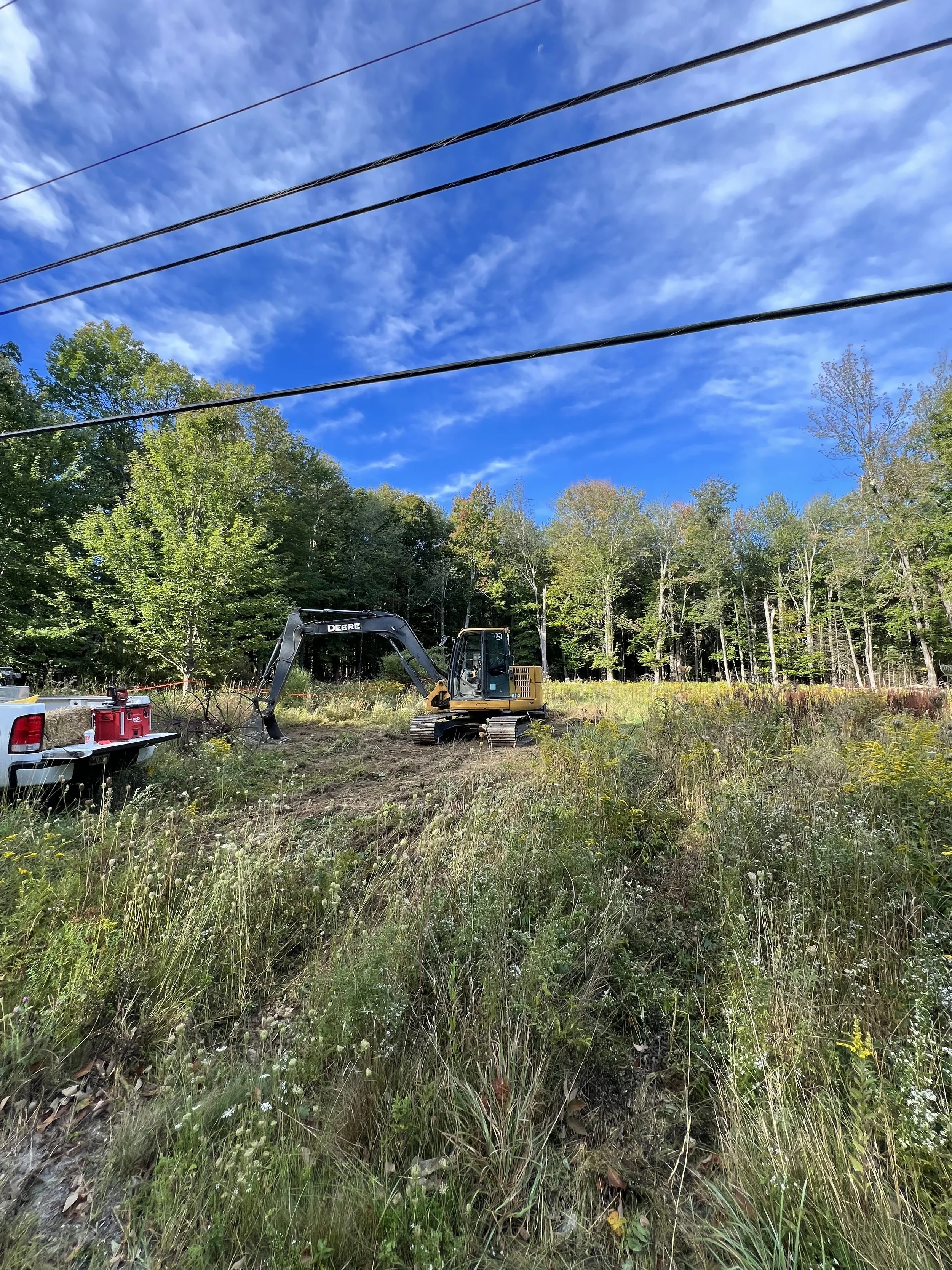 A rural construction site with a small excavator, a truck, and building equipment surrounded by grass and trees, under a partly cloudy blue sky.