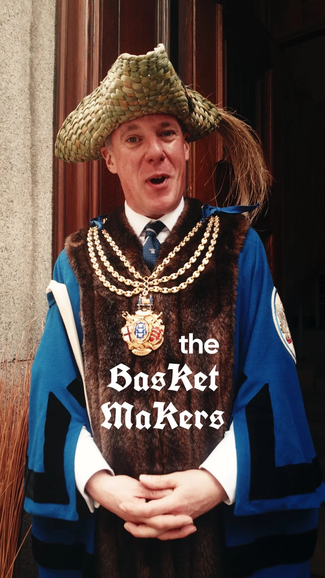 Man dressed in ceremonial Scottish attire, wearing a large woven hat with a feather, and a highland dress with chains and an ornate badge, standing in front of a wooden door.