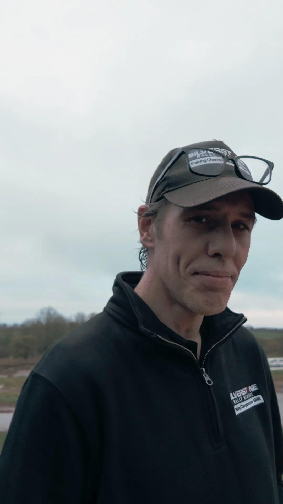 A man outside wearing a black zip-up jacket and a brown cap with glasses resting on top, overcast sky in the background.
