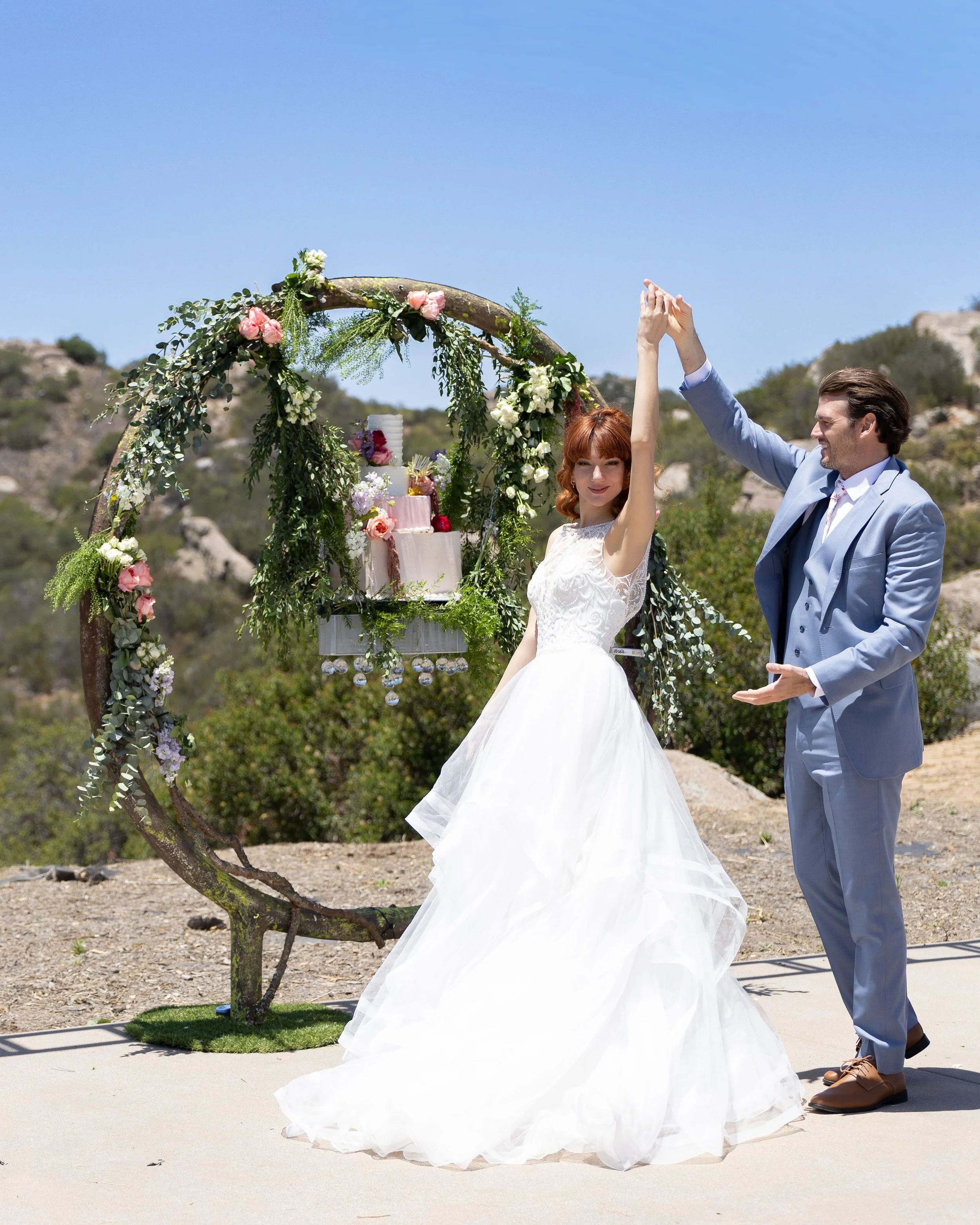 A bride and groom holding hands in an outdoor setting with a floral arch, a wedding cake, and mountains in the background.