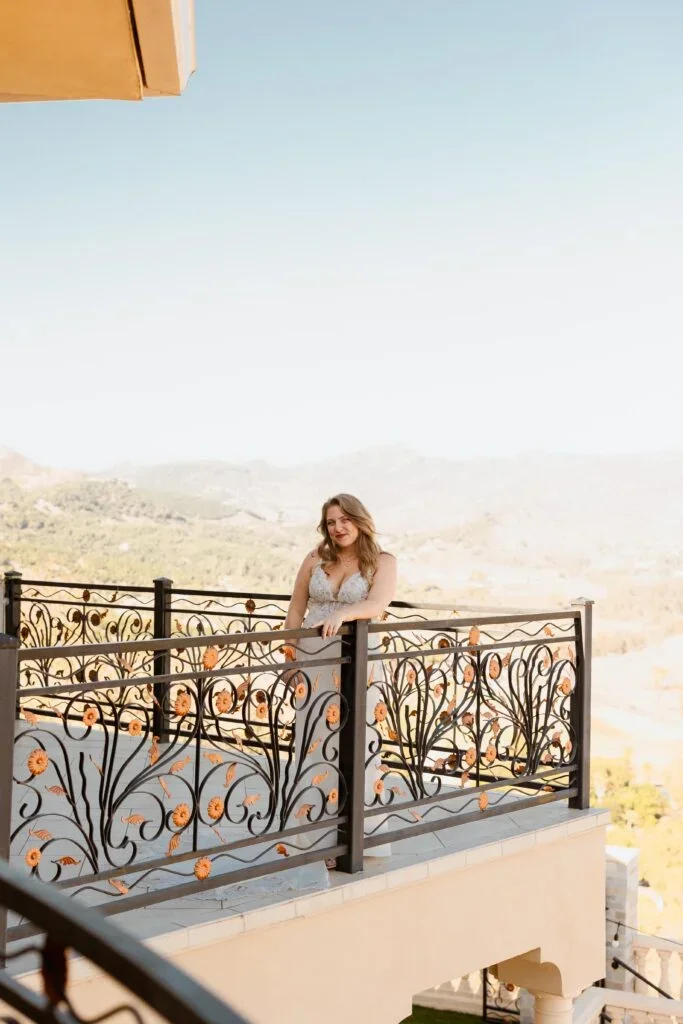 Young woman with wavy hair standing on a balcony with decorative black metal railing with floral design, overlooking a valley with mountains in the background on a sunny day.