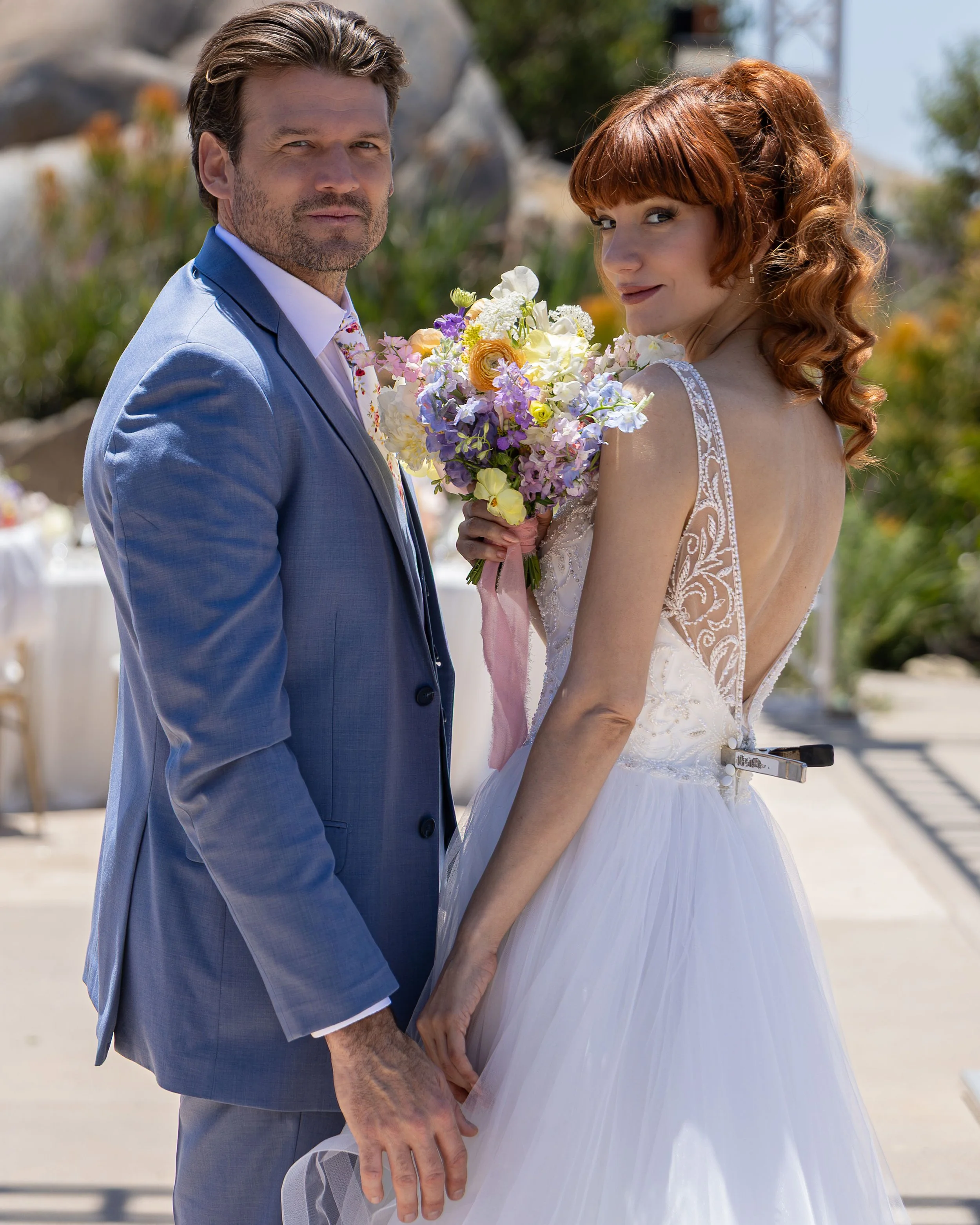 A bride and groom holding hands outdoors on their wedding day. The bride is holding a bouquet of colorful flowers and is dressed in a white wedding gown with lace details. The groom is wearing a light blue suit with a white shirt. They are standing c