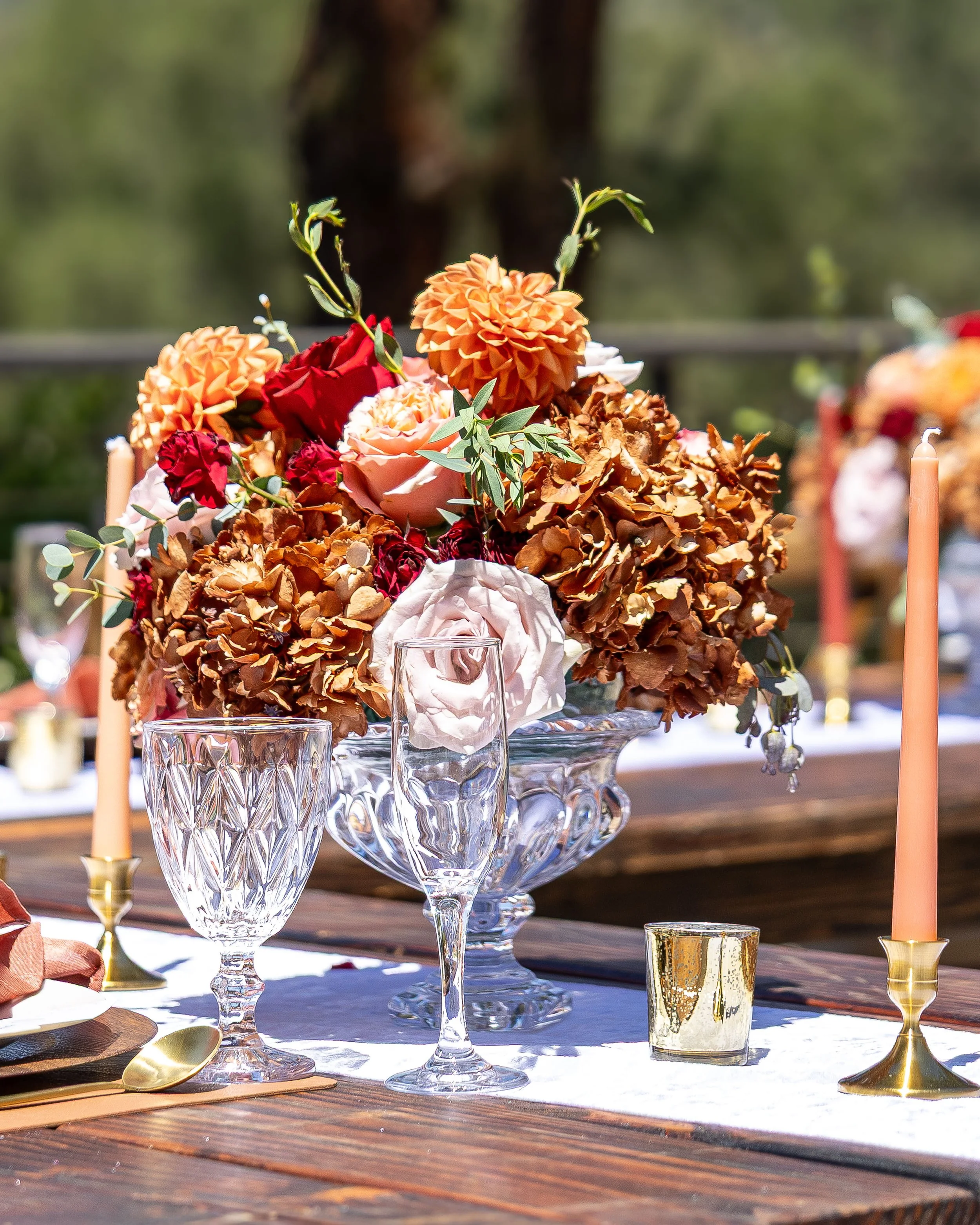A floral table centerpiece consisting of peach, red, and white flowers, set on a wooden table with glassware and candle holders, outdoors with blurred greenery in the background.