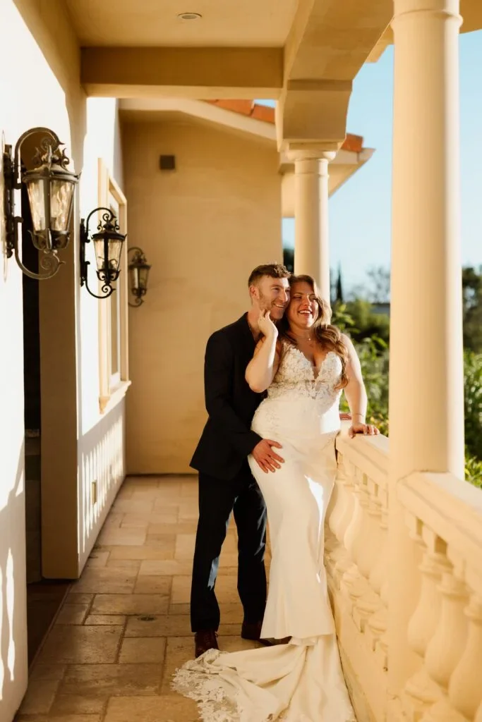 A happy couple in wedding attire standing on a balcony with a decorative railing, looking out and smiling. The woman is wearing a white wedding gown, and the man is in a black suit. The setting appears sunny and elegant.