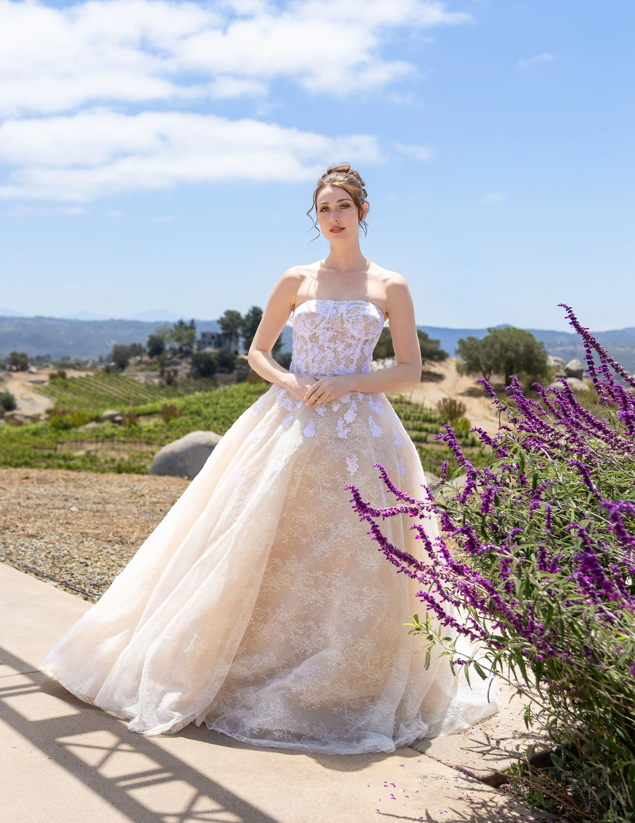 A woman in a white lace wedding dress standing outdoors under a partly cloudy blue sky with a hilly landscape in the background and purple flowers in the foreground.