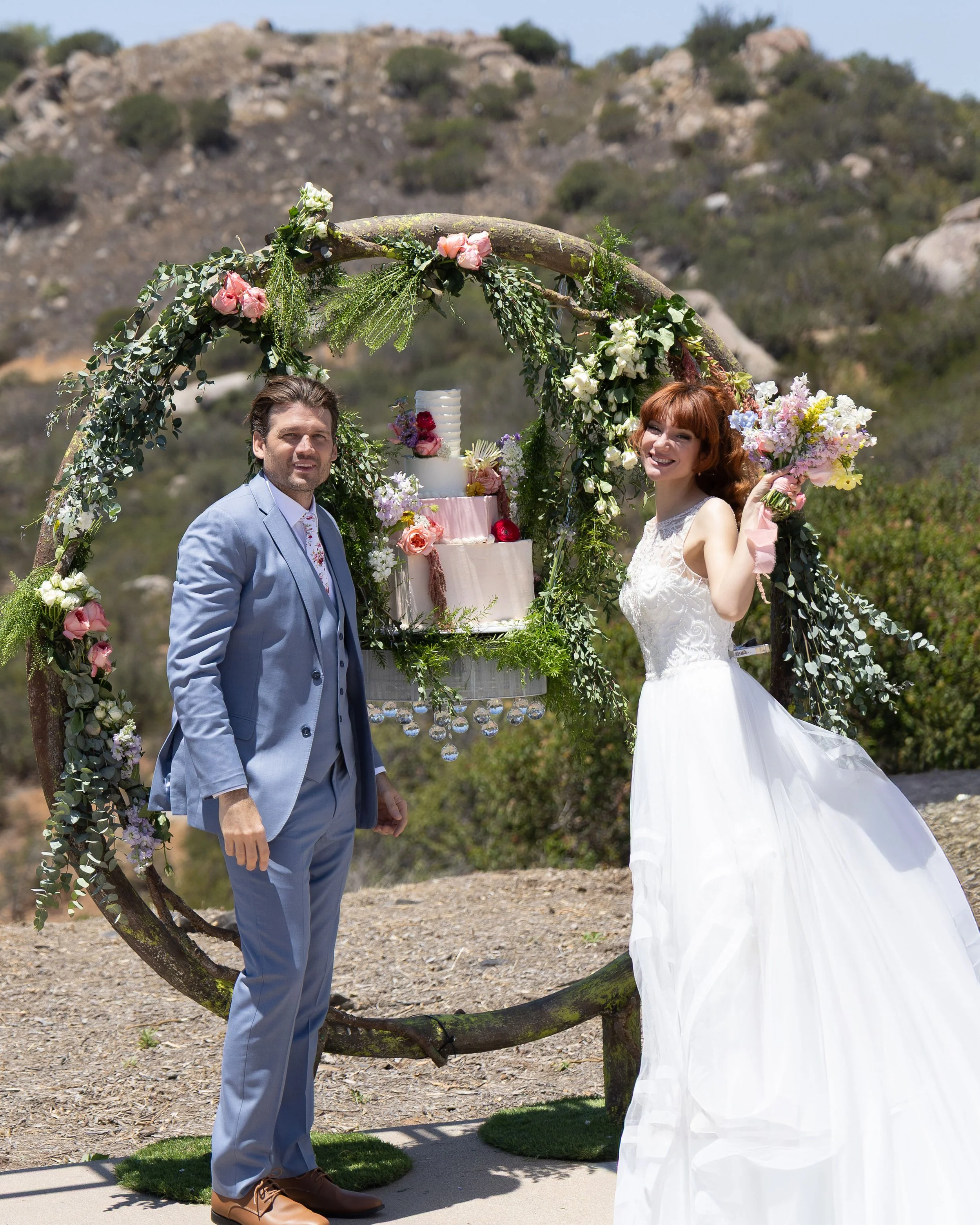 A bride and groom at their outdoor wedding, standing under a decorative arch made of a tree branch wrapped in flowers and greenery, with a wedding cake on a stand behind them, set in a desert landscape with hills and rocks in the background.