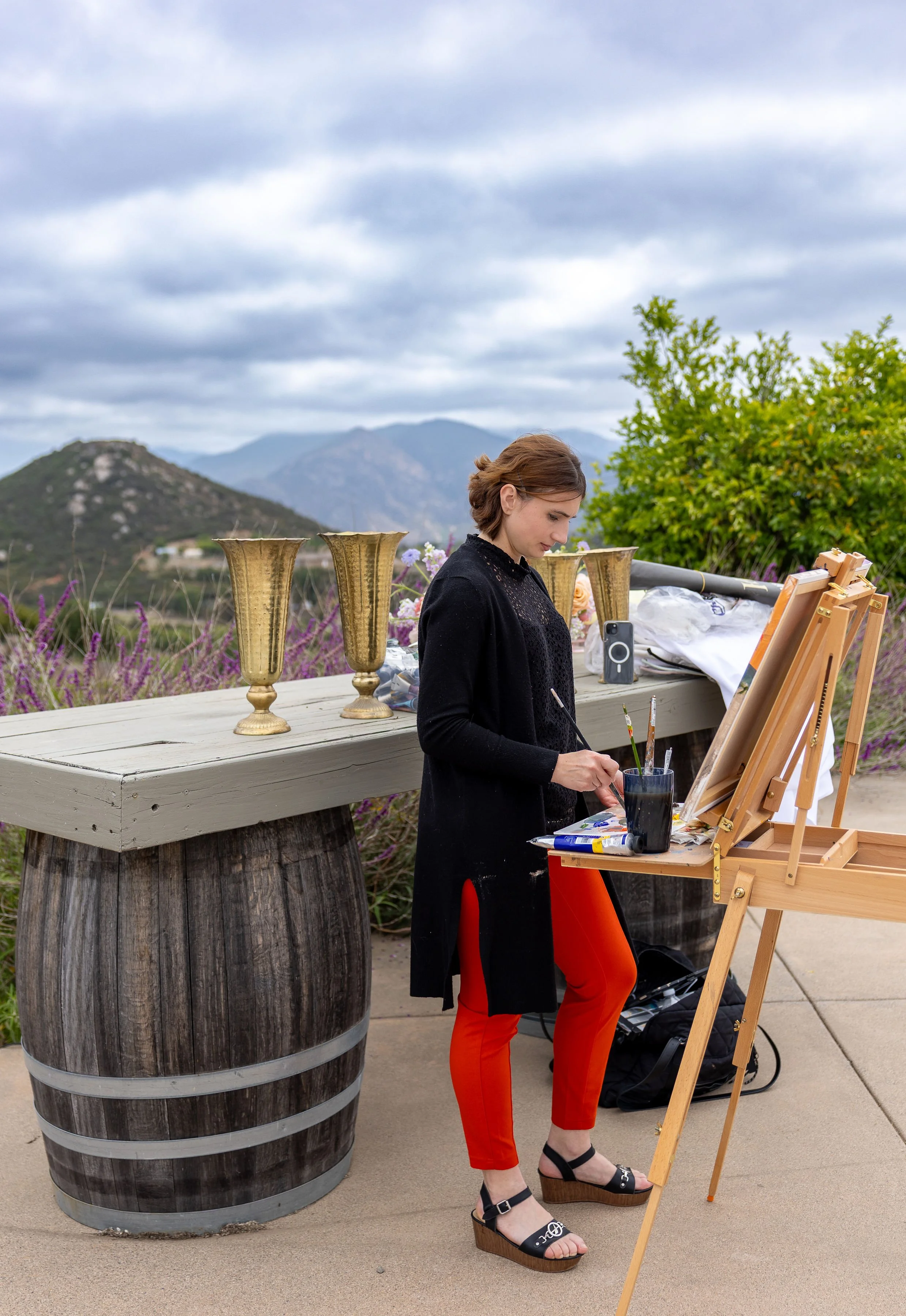 A woman with short brown hair wearing a black top and red pants with black wedge sandals painting on an easel outdoors, with mountains in the background, a large wooden barrel, and three brass goblets on a table nearby.