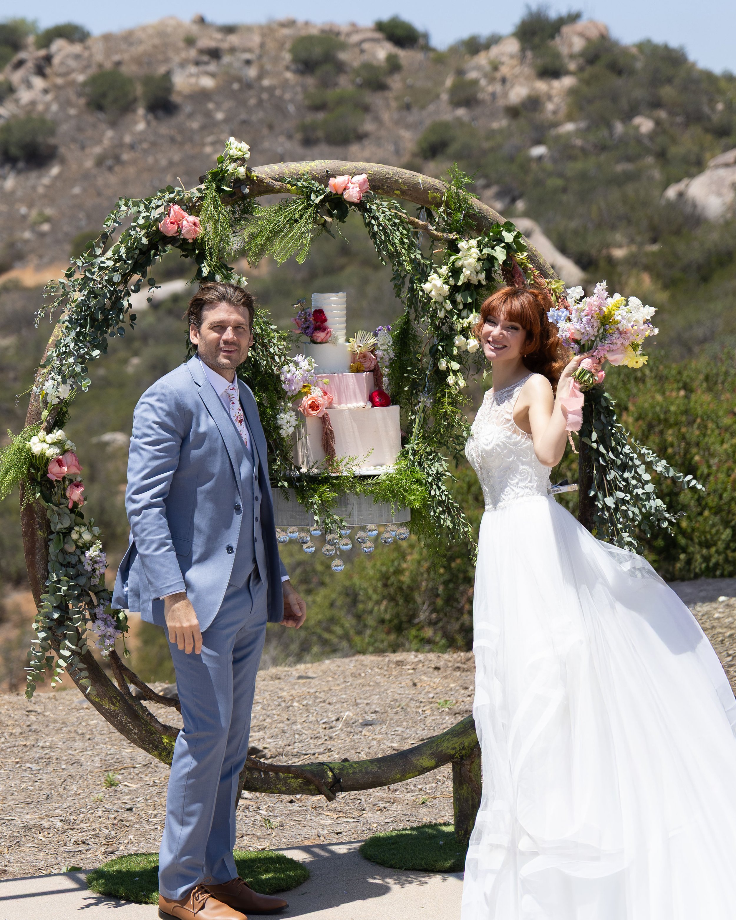 A bride and groom standing outdoors near a decorated arch with flowers and a tiered wedding cake, with rocky hills in the background.