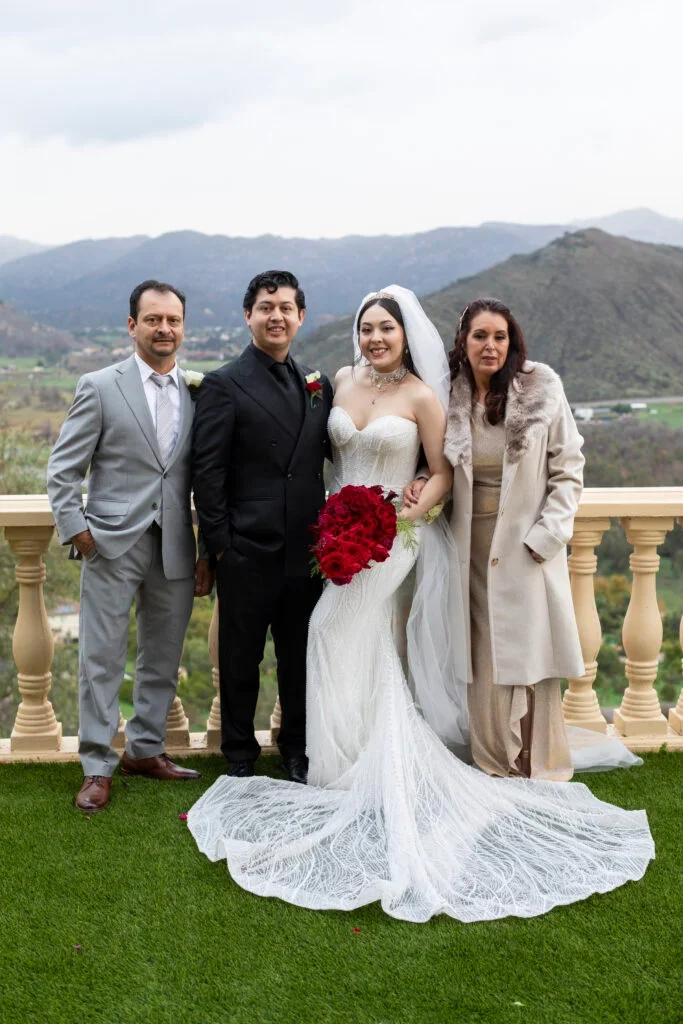 Wedding photo outdoors featuring a bride in a white gown holding a bouquet of red roses, groom in a black suit, and two other adults dressed elegantly, with mountains in the background.