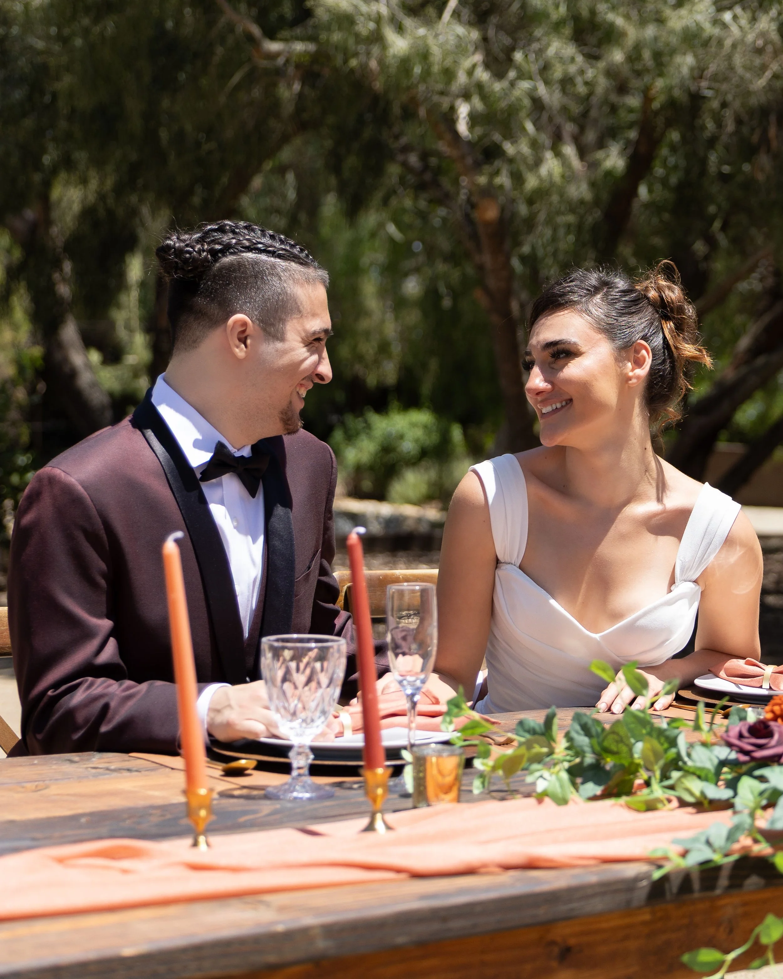 A smiling man and woman are sitting at a table outdoors, engaged with each other, with glasses, candles, and greenery on the table.
