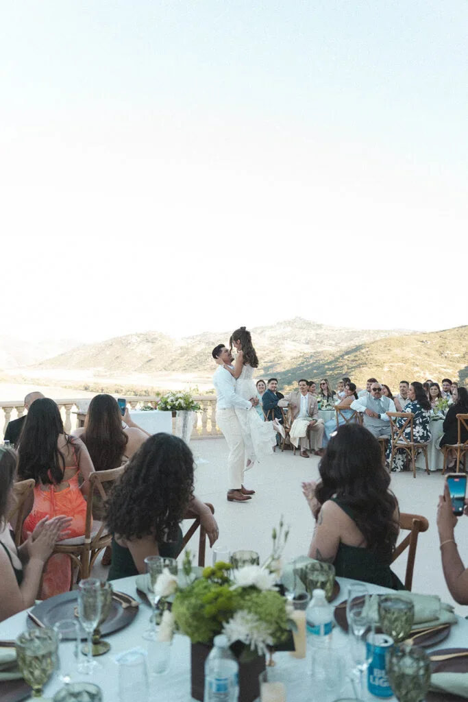 A couple in wedding attire sharing a dance at an outdoor wedding reception with guests seated at decorated tables and mountain scenery in the background.