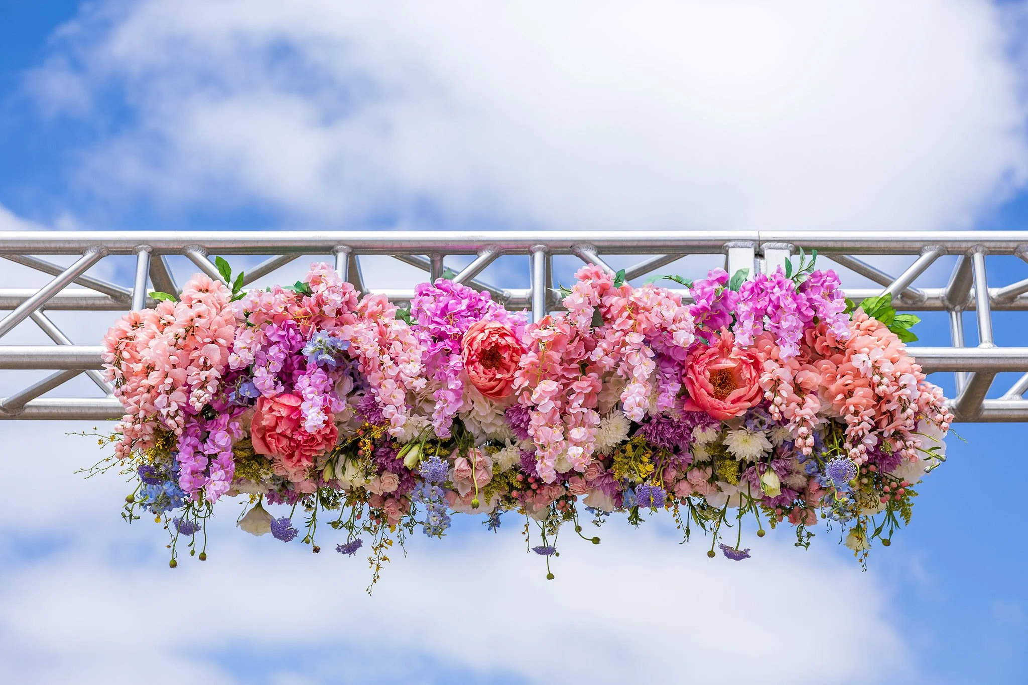 A floral arrangement of pink, purple, and peach flowers hanging under a metal truss against a partly cloudy blue sky.