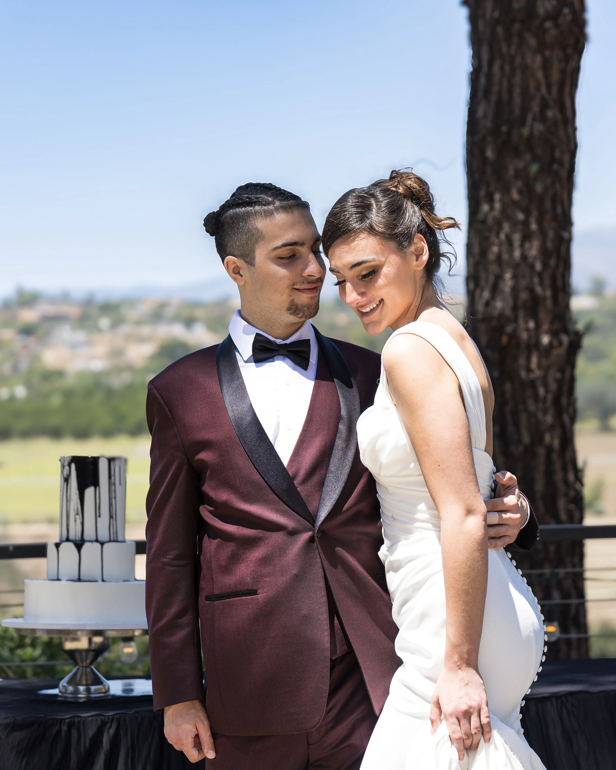 A couple dressed in formal attire, standing outdoors near a tree, sharing a tender moment with eyes closed and heads touching. A multi-tiered wedding cake is in the background on a table.