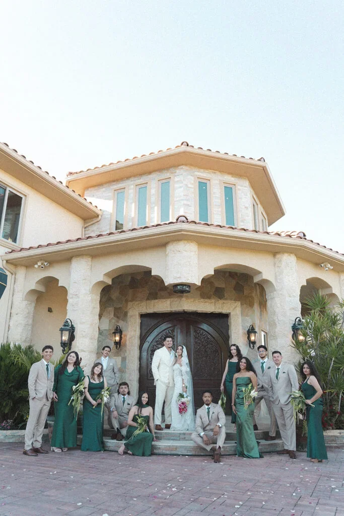 A wedding group standing in front of a large house with a stone facade and arched entryway, including a bride and groom in the center, surrounded by bridesmaids and groomsmen.