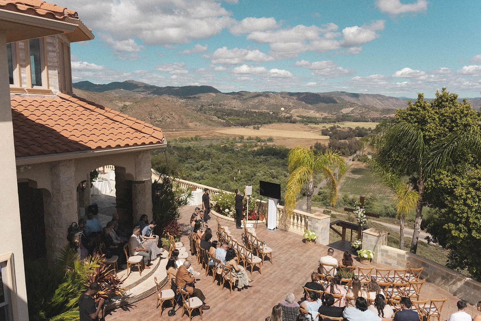 Outdoor wedding ceremony with guests seated in rows, a pastor or officiant at the front, and scenic hillside landscape with trees, mountains, sky, and clouds in the background.